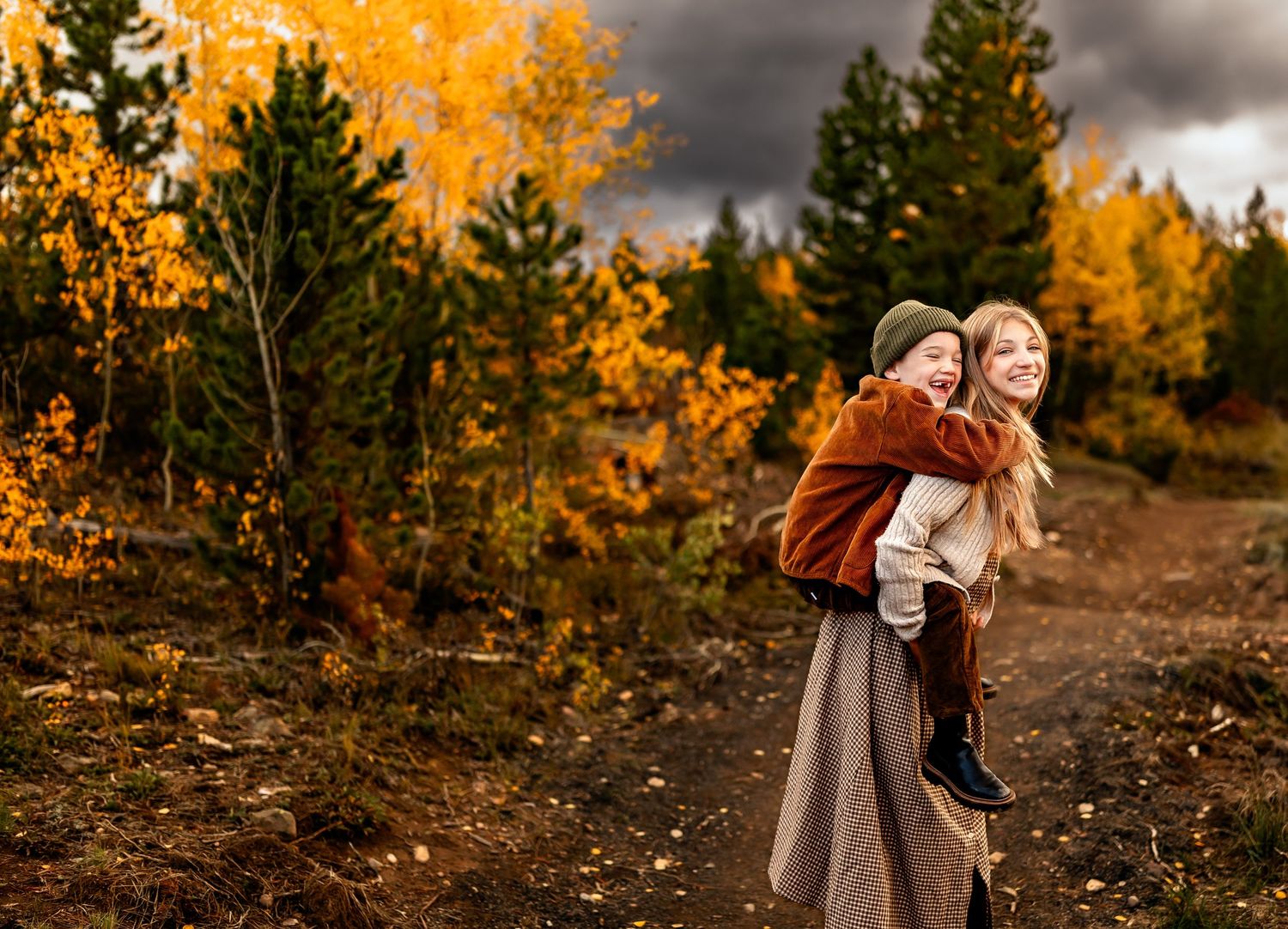 A smiling girl gives a piggyback ride to a younger child amid vibrant autumn foliage on a cloudy day.