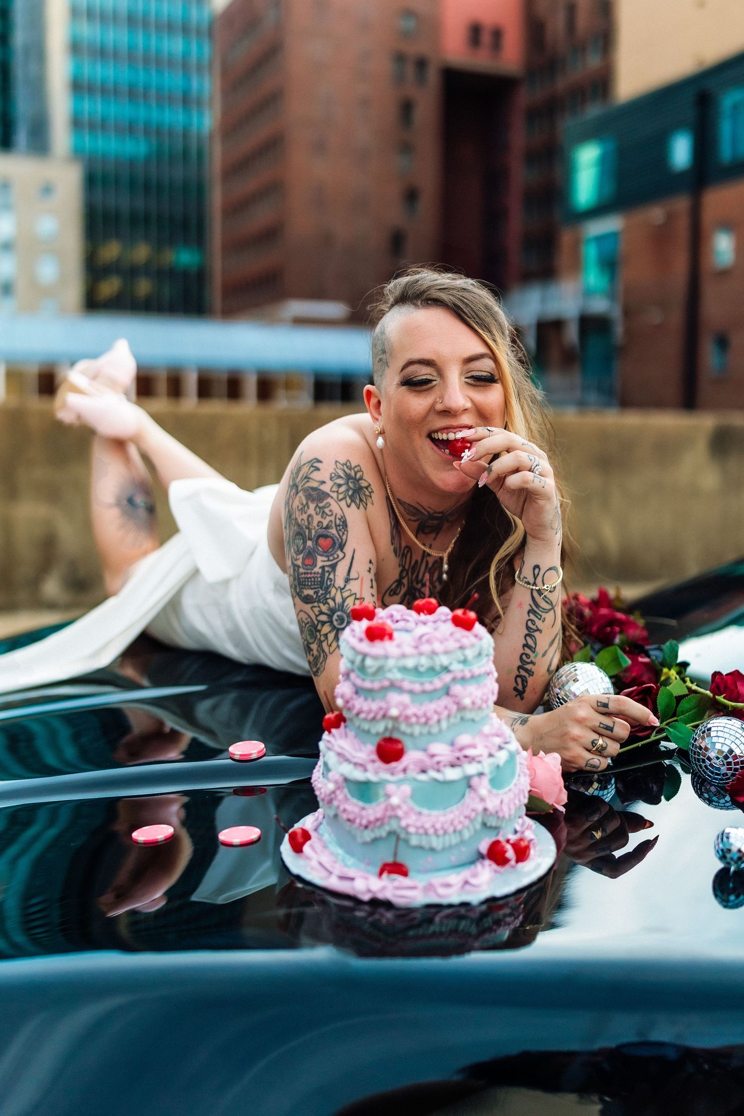 Tattooed woman in white dress lying on car hood eating cake with cherries, urban rooftop setting.