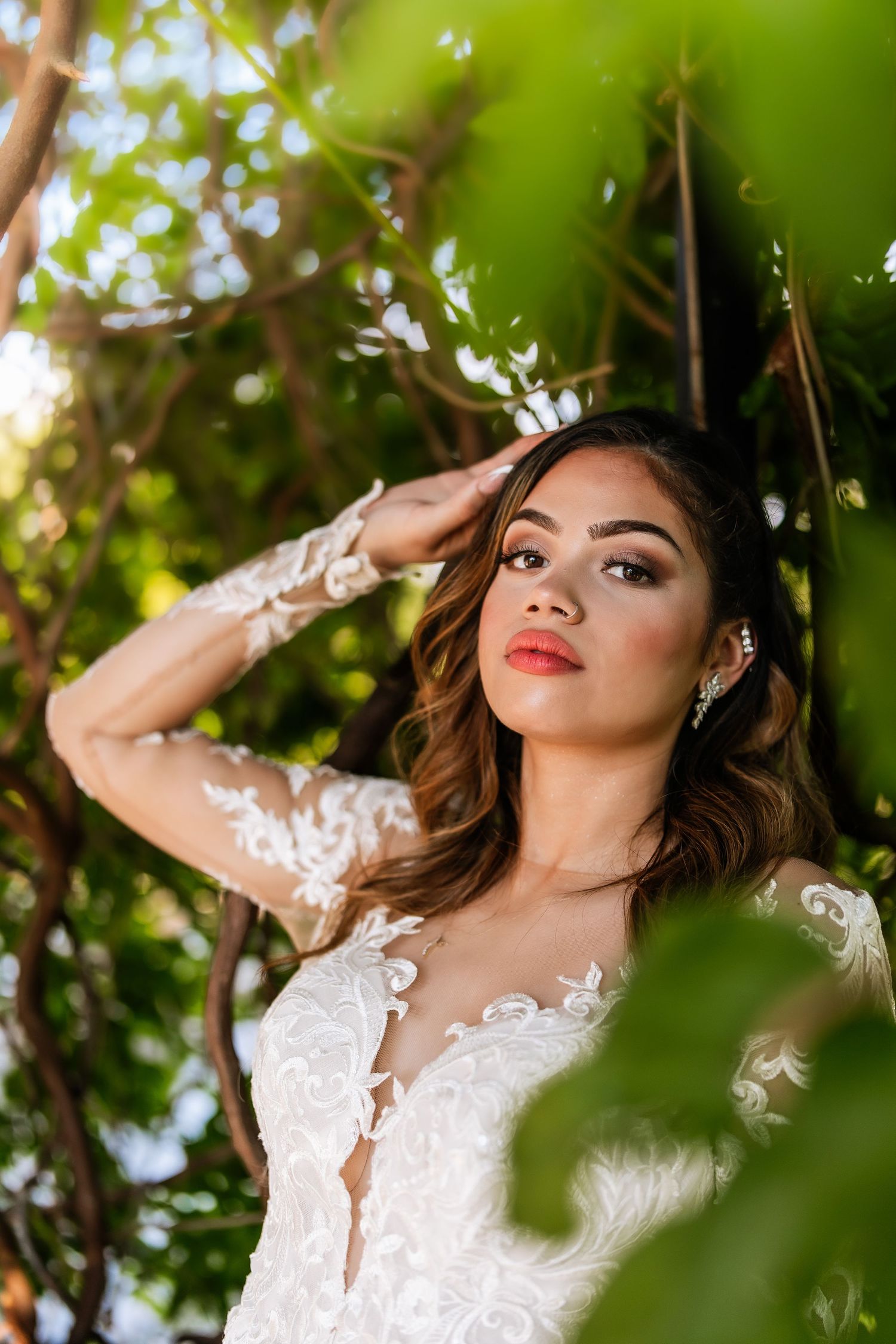 Bride in lace long-sleeve wedding gown poses among green trees, showcasing elegant bridal makeup and earrings.