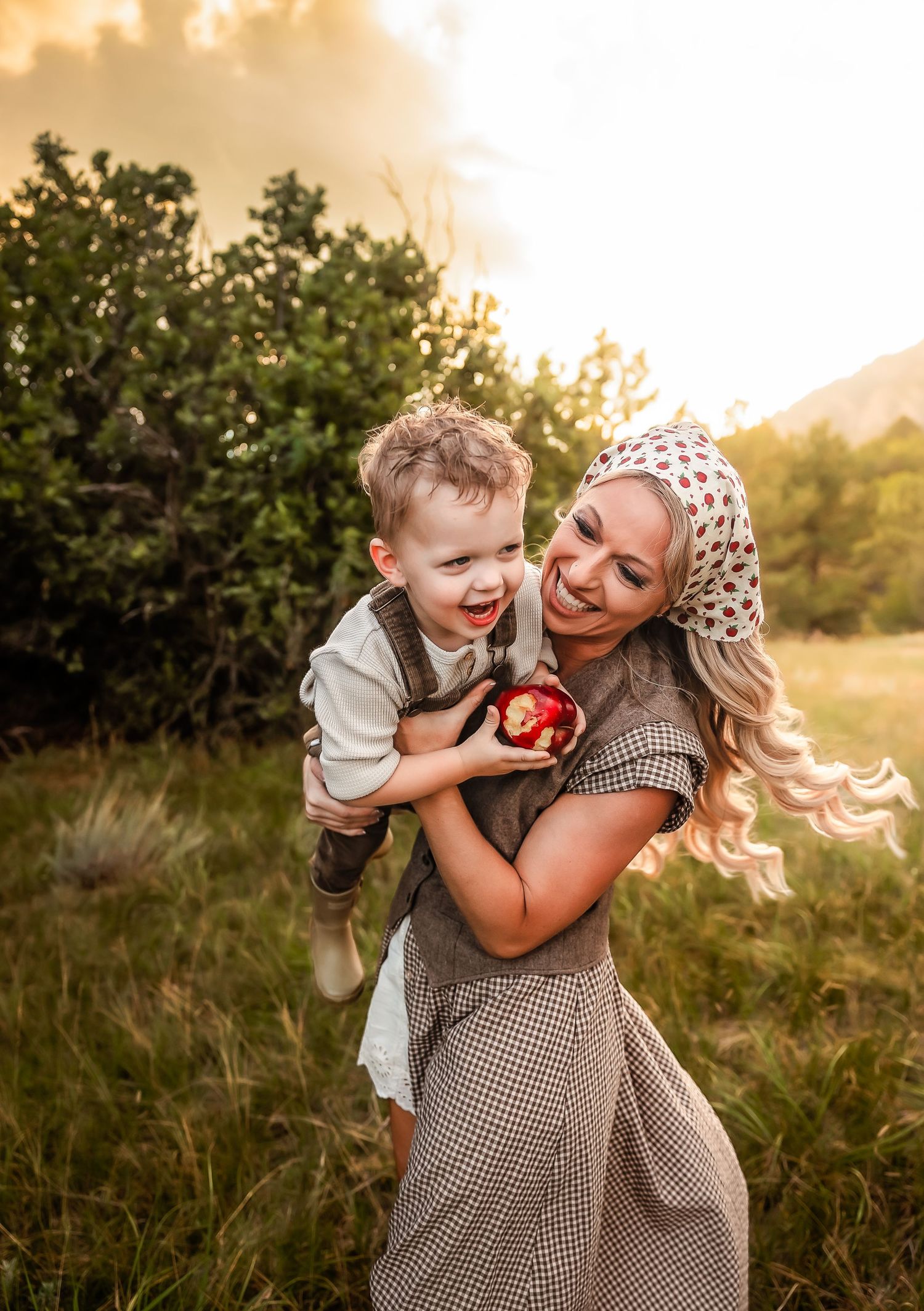 Smiling woman in a checkered dress and floral headscarf lifts a laughing toddler holding a red apple in a golden sunset field.