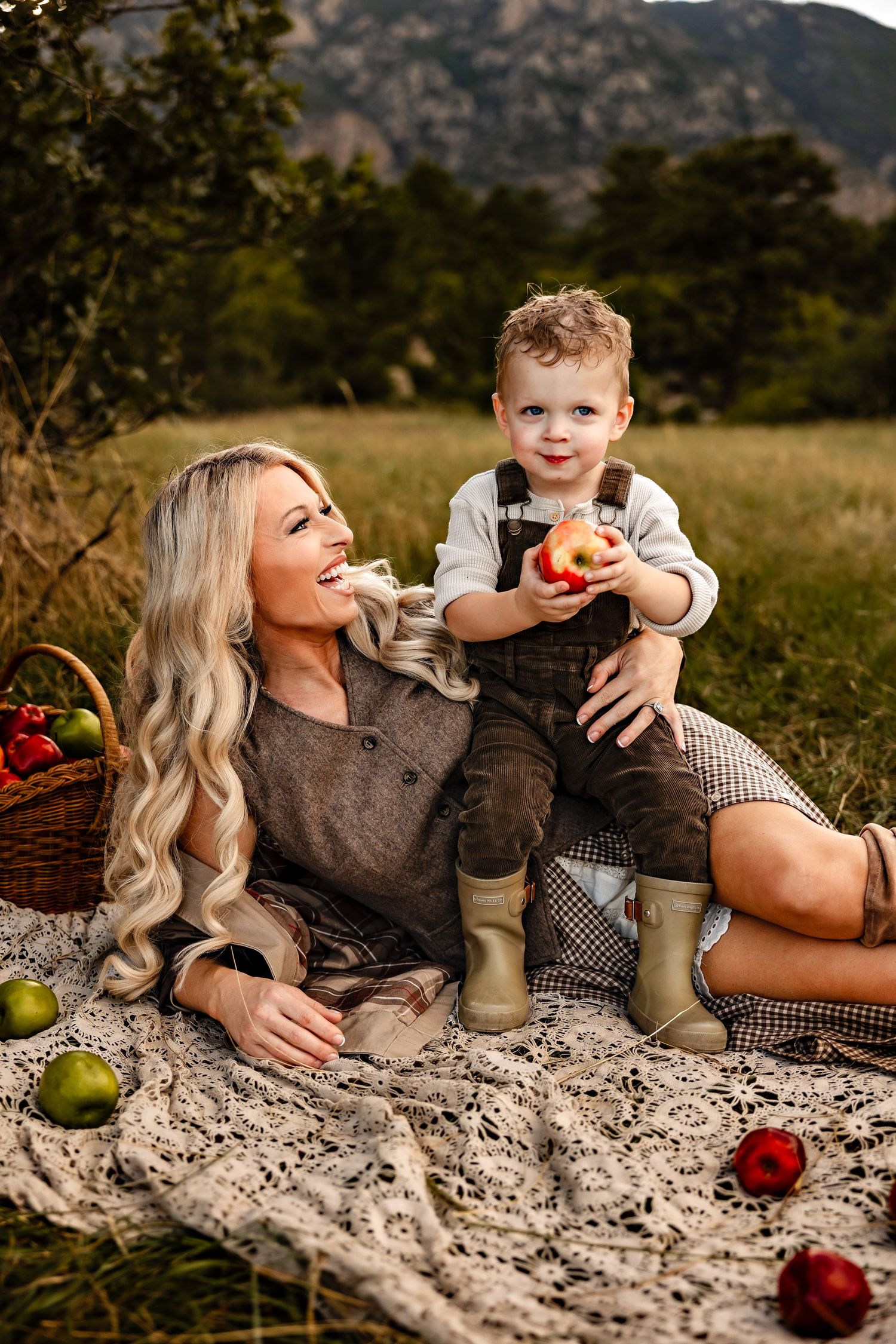A smiling blonde woman reclines on a lace blanket with her toddler son holding a red apple, with apples nearby and mountains behind.