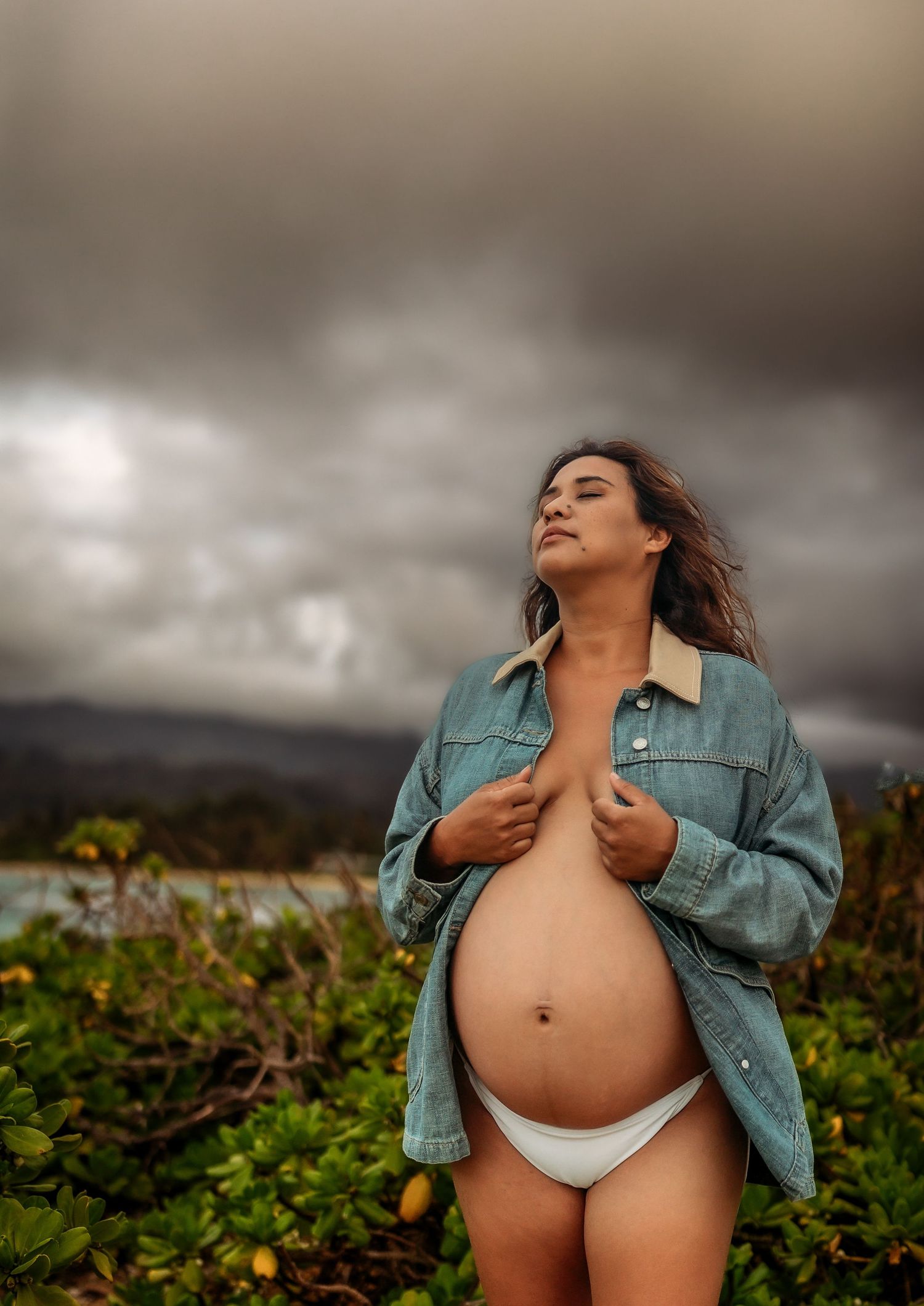 Pregnant woman in denim jacket and white bikini bottom poses outdoors under dramatic cloudy sky near lush greenery.