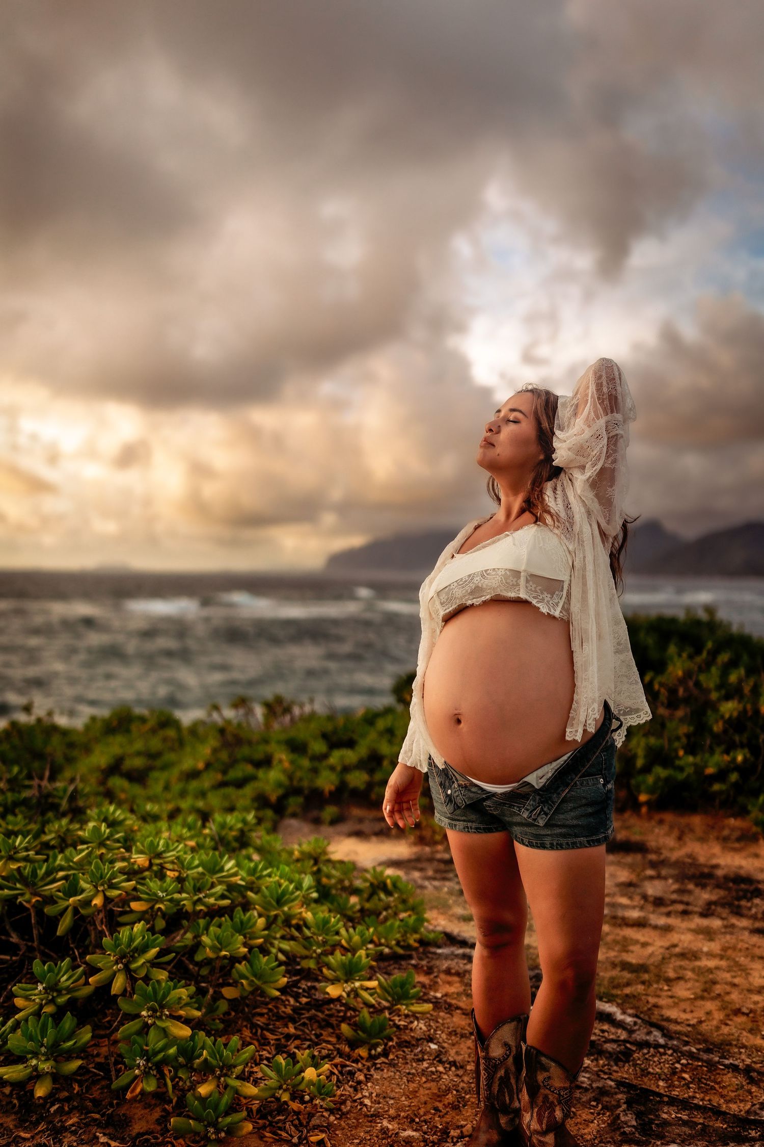 Pregnant woman in white lace top and denim shorts poses on Hawaii coastline at dramatic sunset.