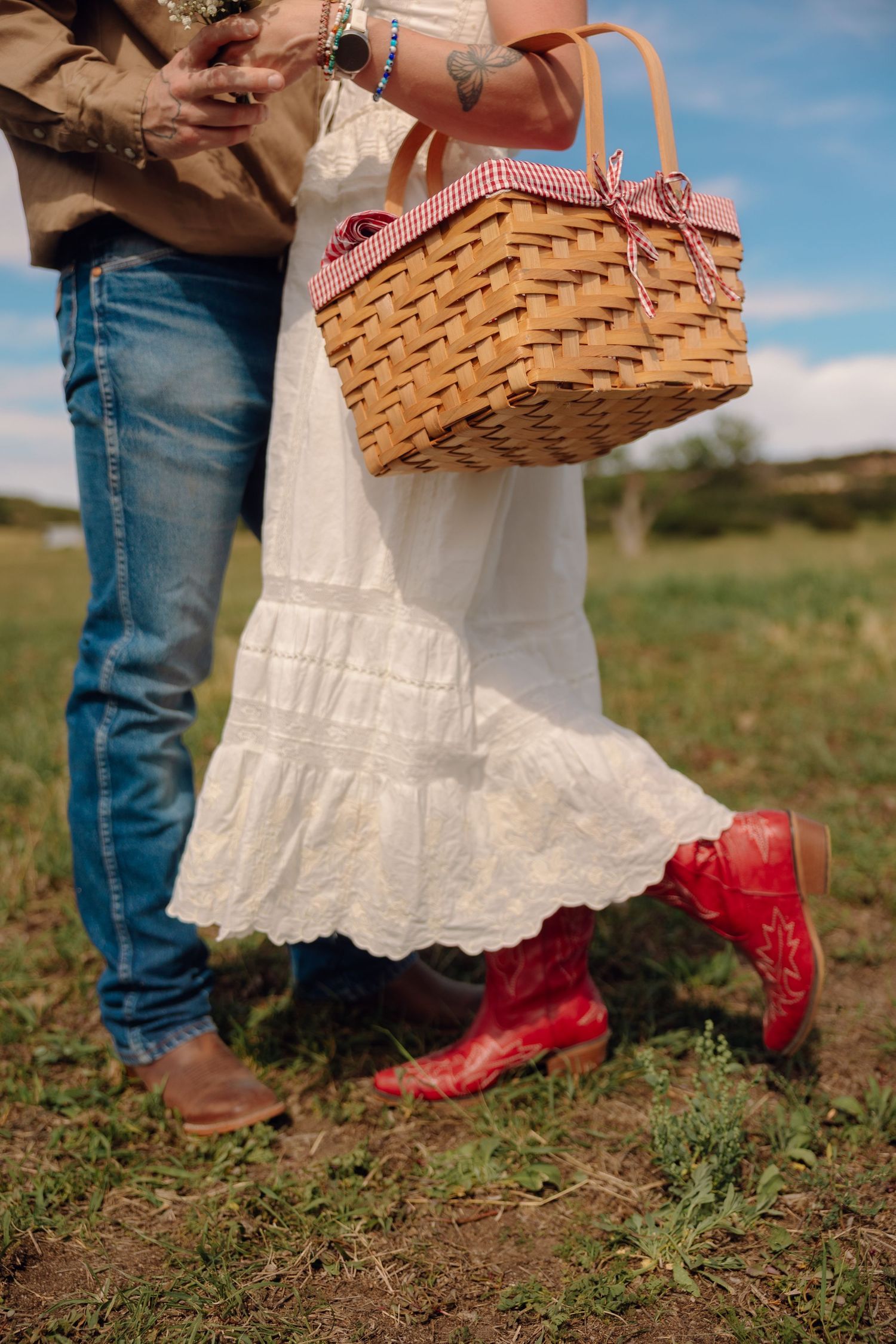 Couple in white dress and red cowboy boots holding a wicker picnic basket in a green field.