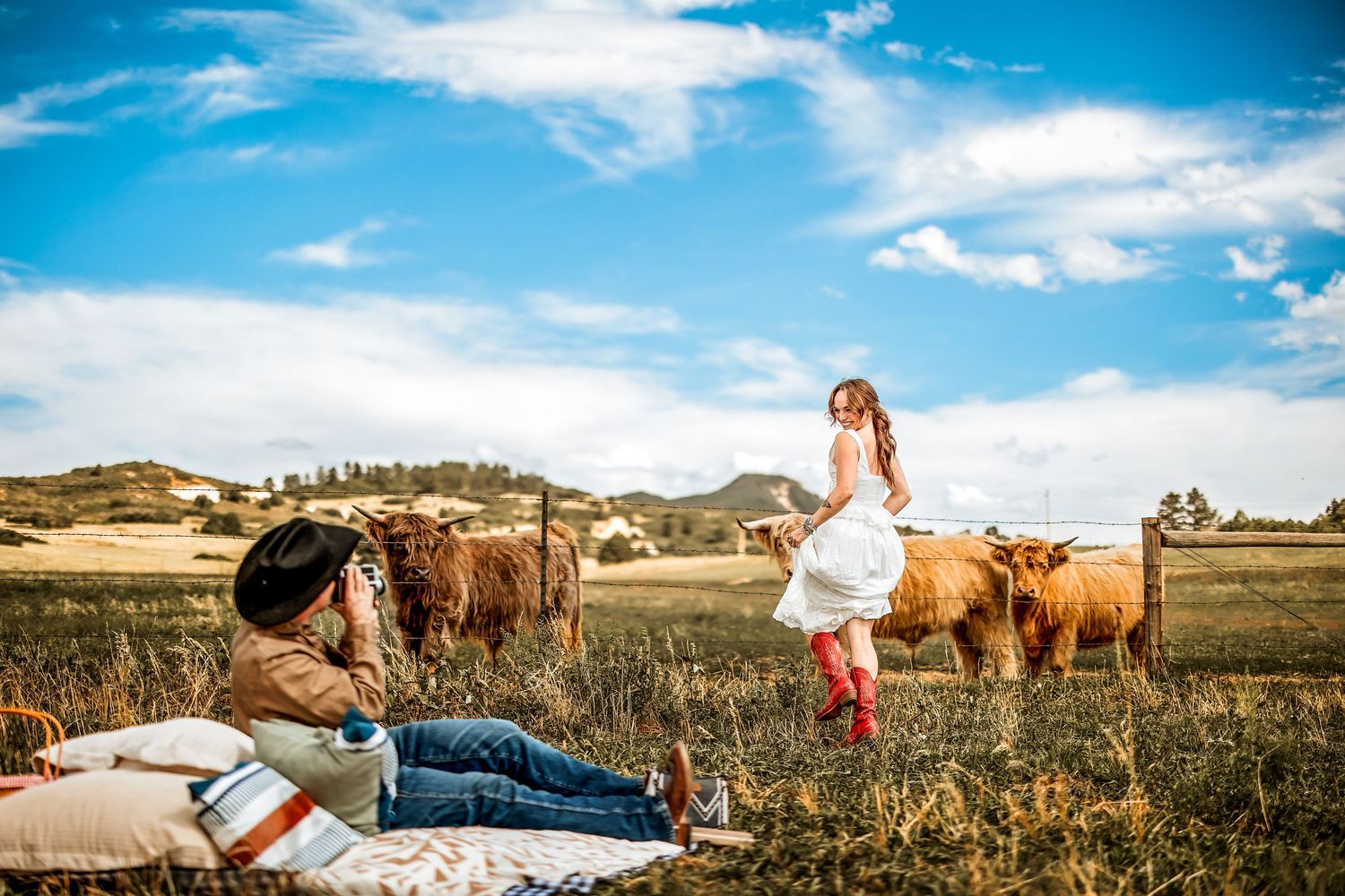 Photographer lying on blanket capturing woman in white dress posing with Highland cattle in scenic field under blue sky.