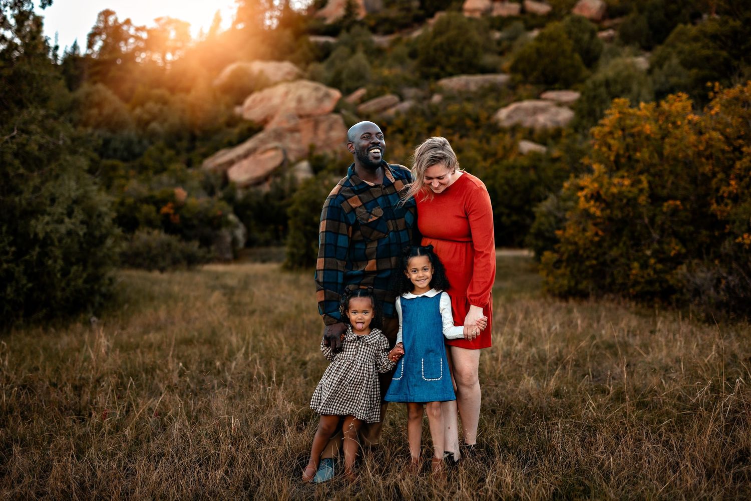 Family of four, two parents and two young girls, pose together in a grassy field with rock formations and trees in golden sunset light.