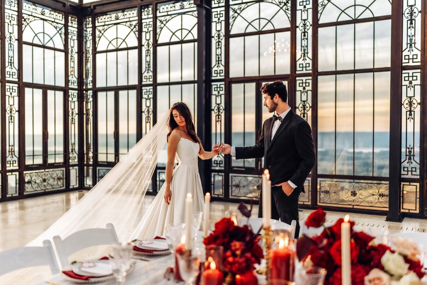 Elegant bride and groom hold hands in a stunning glass-walled venue with red floral table decor and panoramic views.