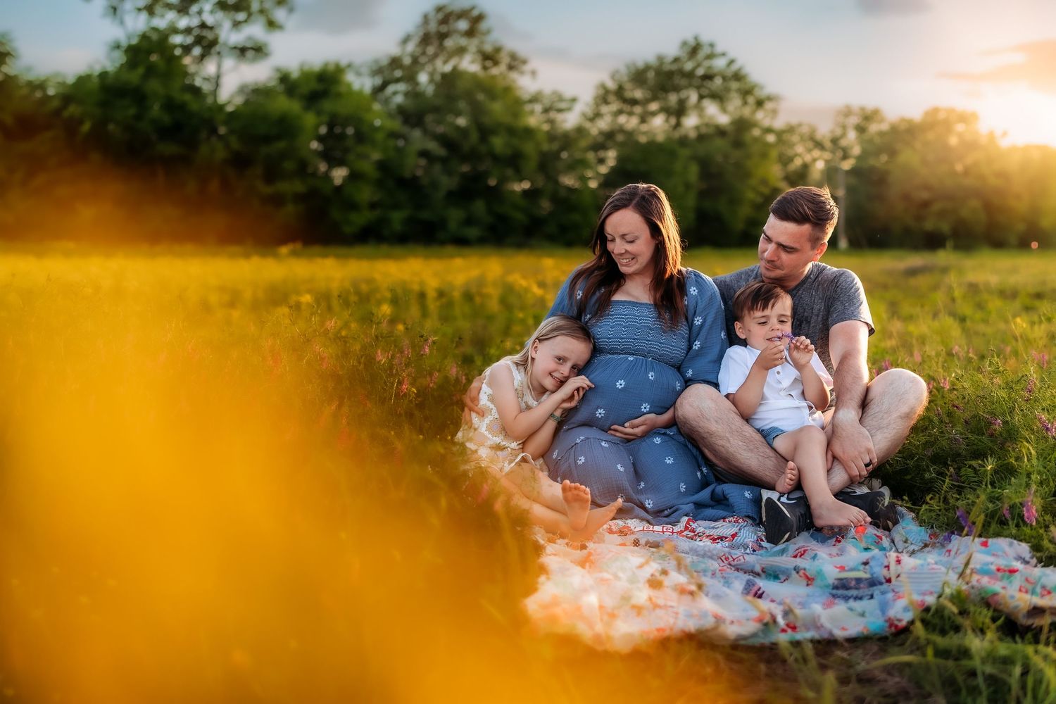 Family maternity photo session outdoors at golden hour, parents and two children sitting on blanket in sunny green field.