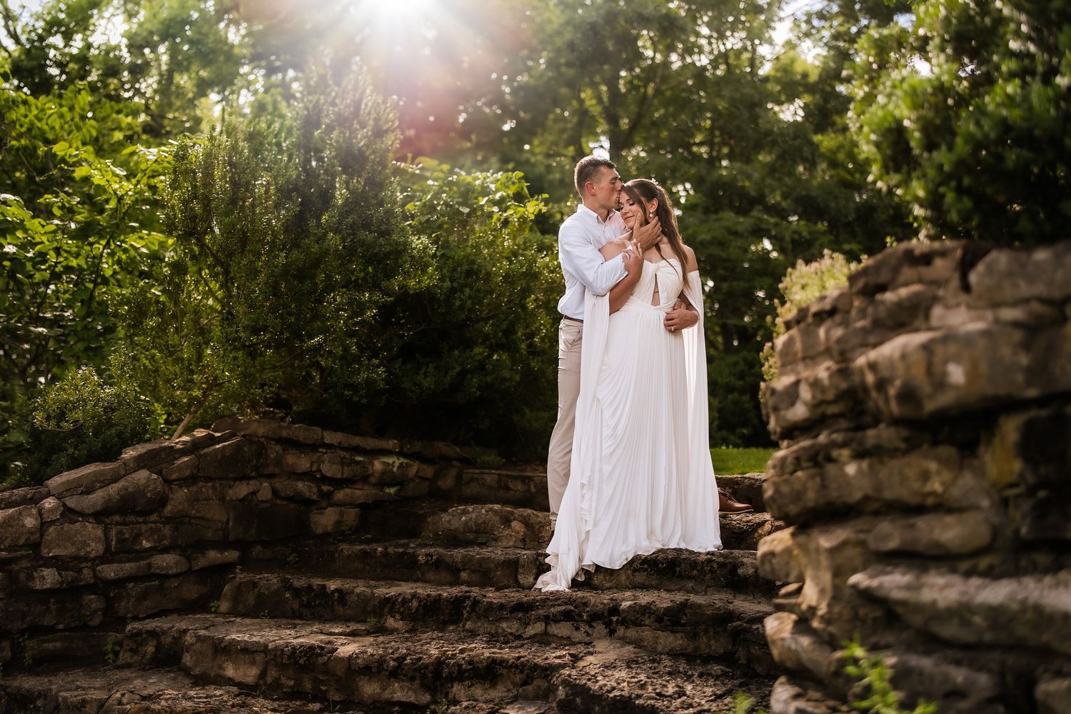 Bride and groom embrace on stone steps surrounded by lush greenery in golden sunlight during outdoor wedding.
