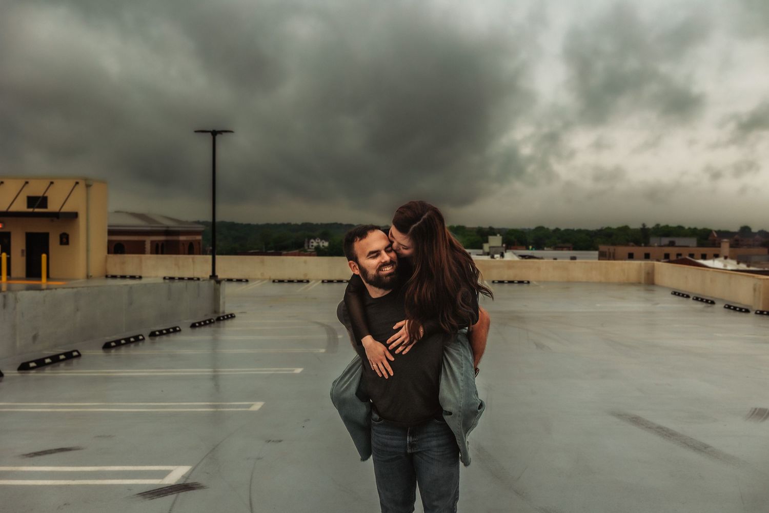 Couple sharing a piggyback ride on rooftop parking lot under dramatic stormy sky.