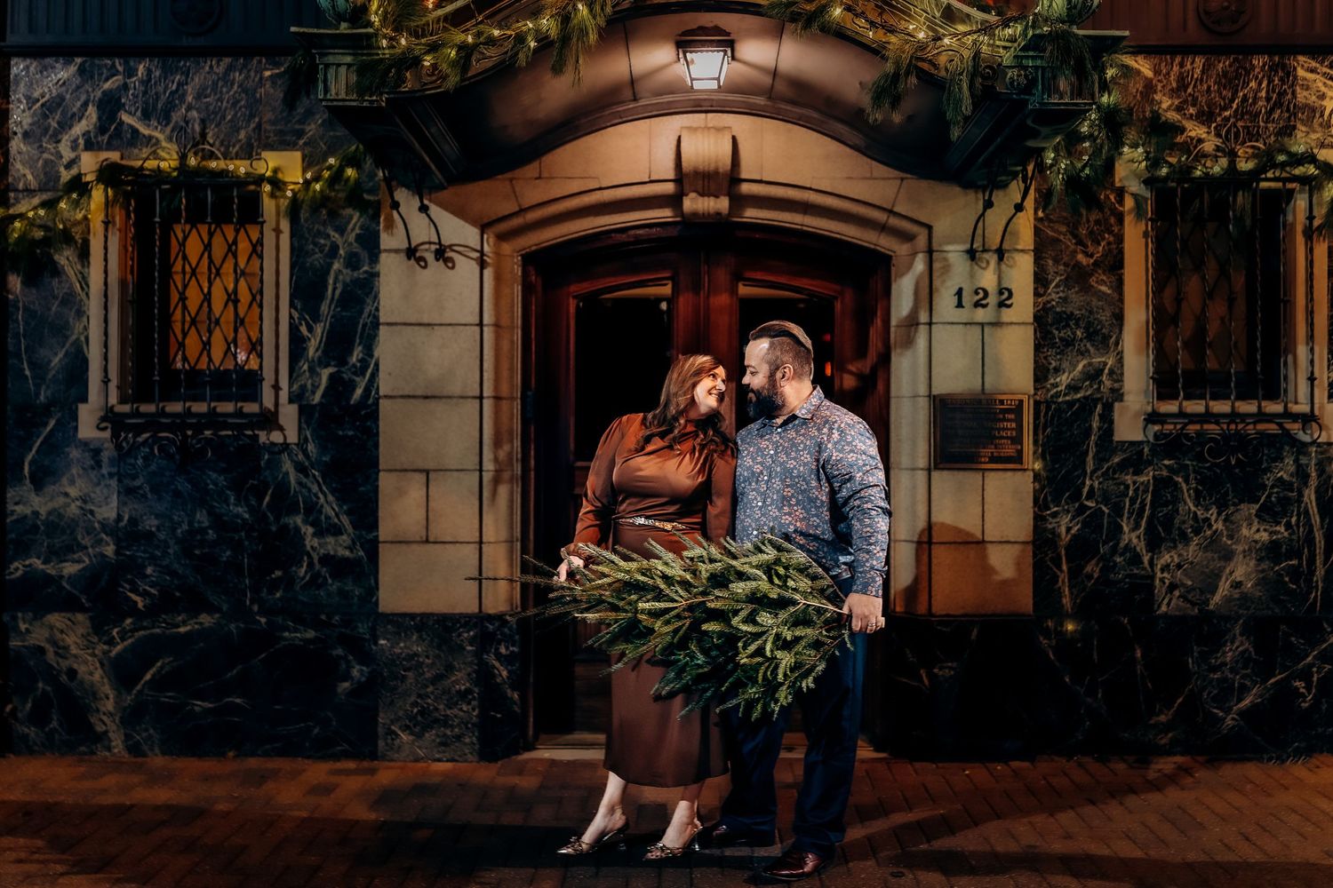 Couple holding greenery bouquet standing outside historic building entrance at night during holiday season.