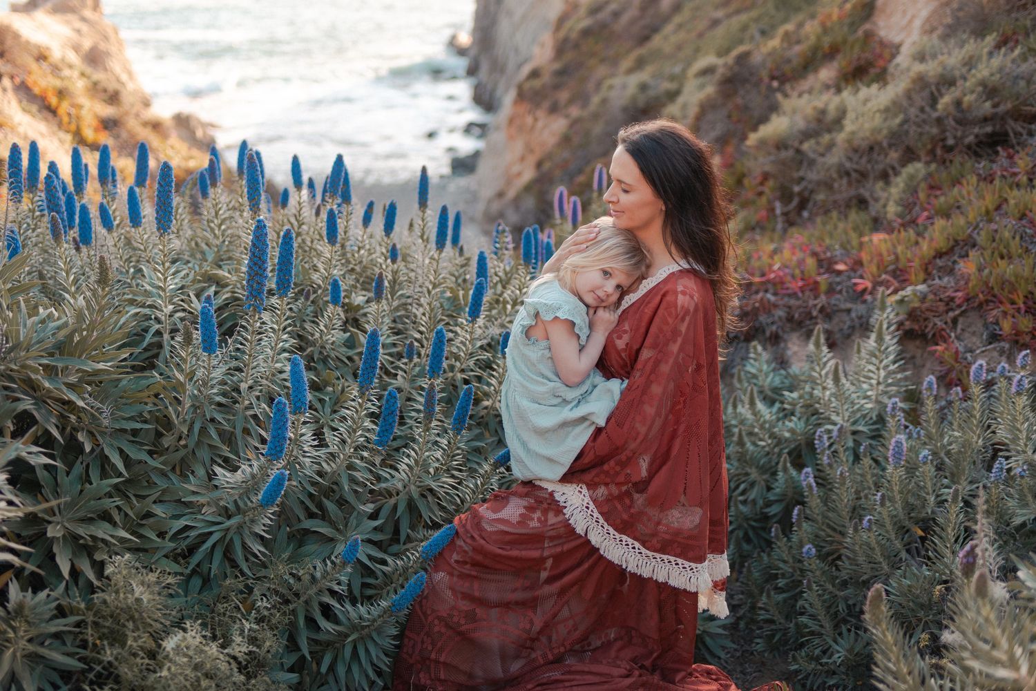 Mother in red dress holds baby among blue wildflowers on a coastal cliffside at golden hour.