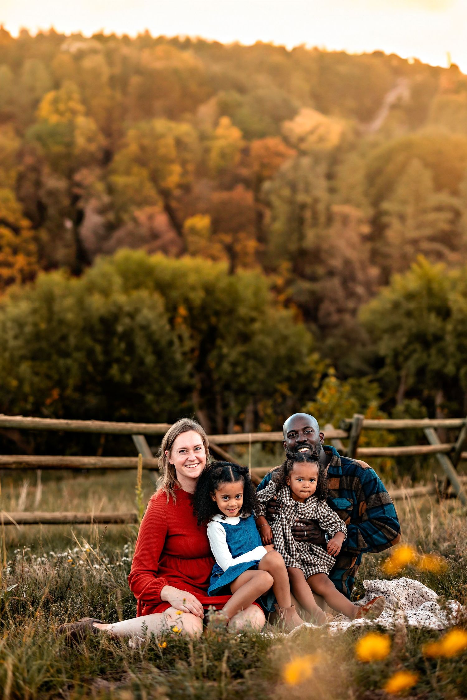 Smiling family of four sitting together on a grassy hillside with yellow wildflowers, wooden fence, and colorful autumn trees behind them.