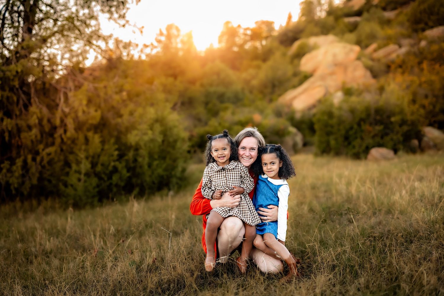 A smiling woman kneels in a grassy field hugging two young girls, with golden sunset light and rocky hills glowing in the background.