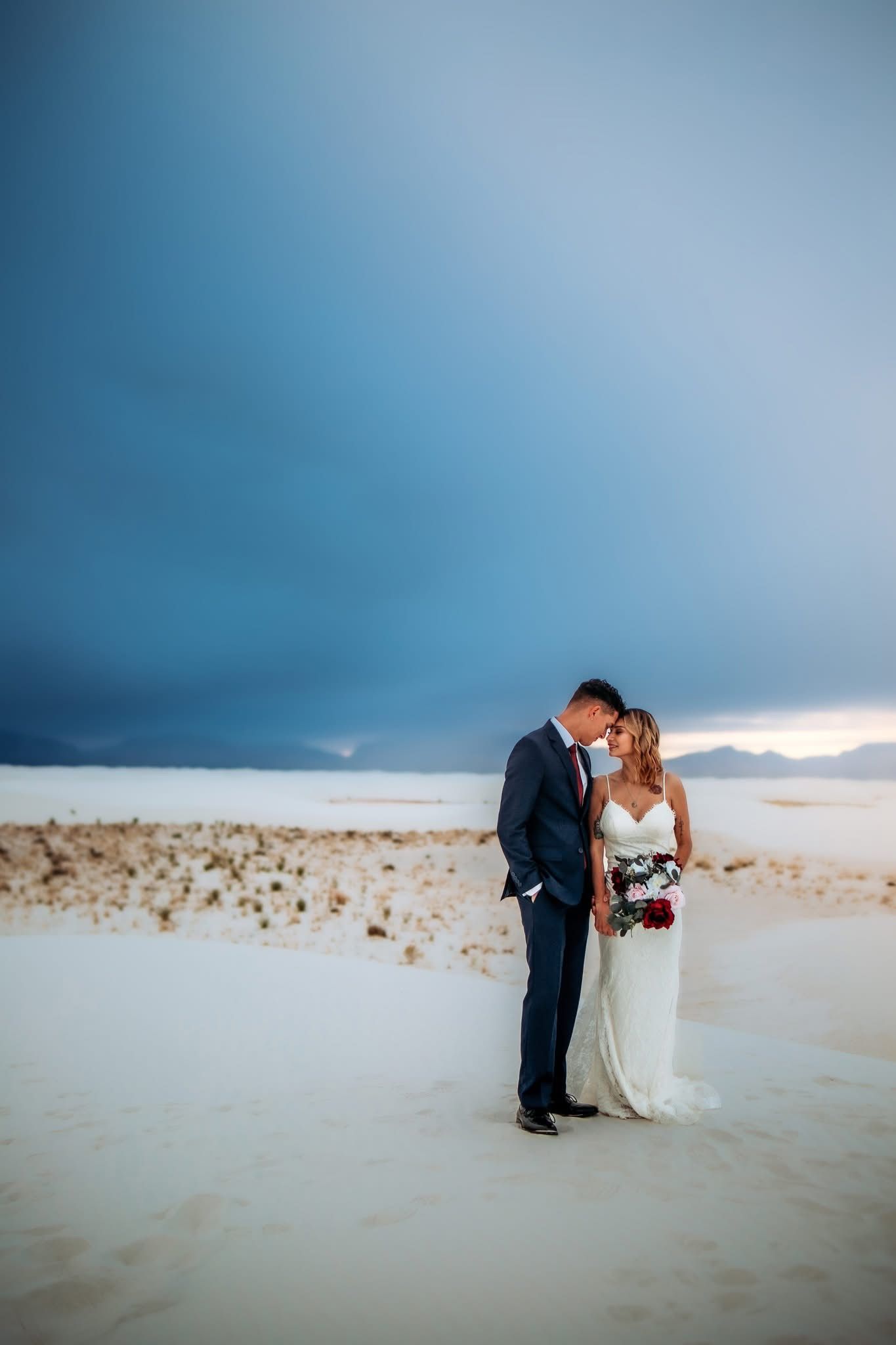 Wedding couple touches foreheads tenderly on white sand dunes under a dramatic stormy sky.