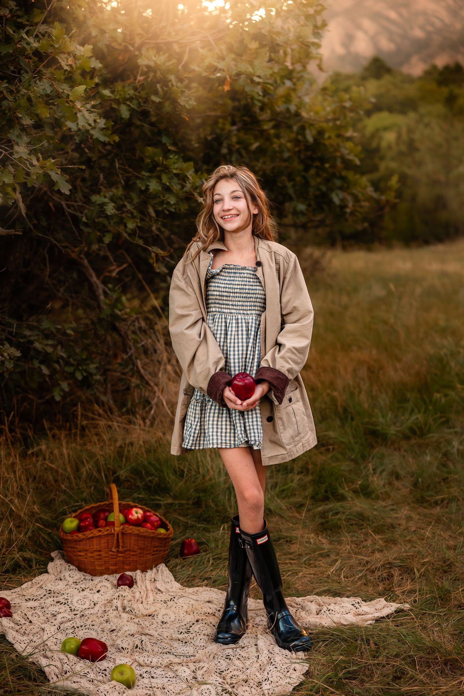 A smiling girl in a plaid dress, trench coat, and rain boots holds a red apple near a wicker basket at golden hour.