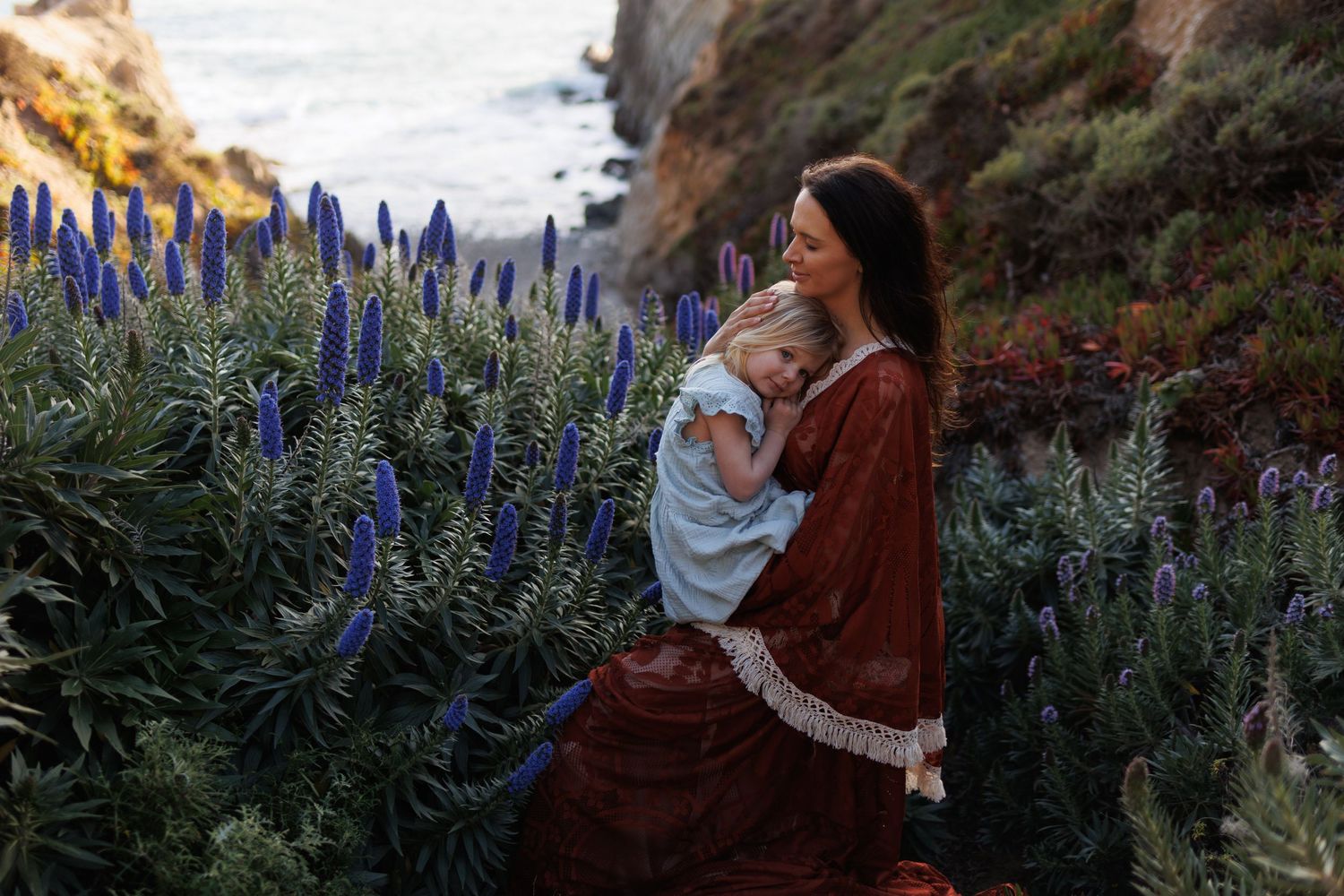 A mother in a red dress cradles her young child among tall blue wildflowers on a coastal cliffside.