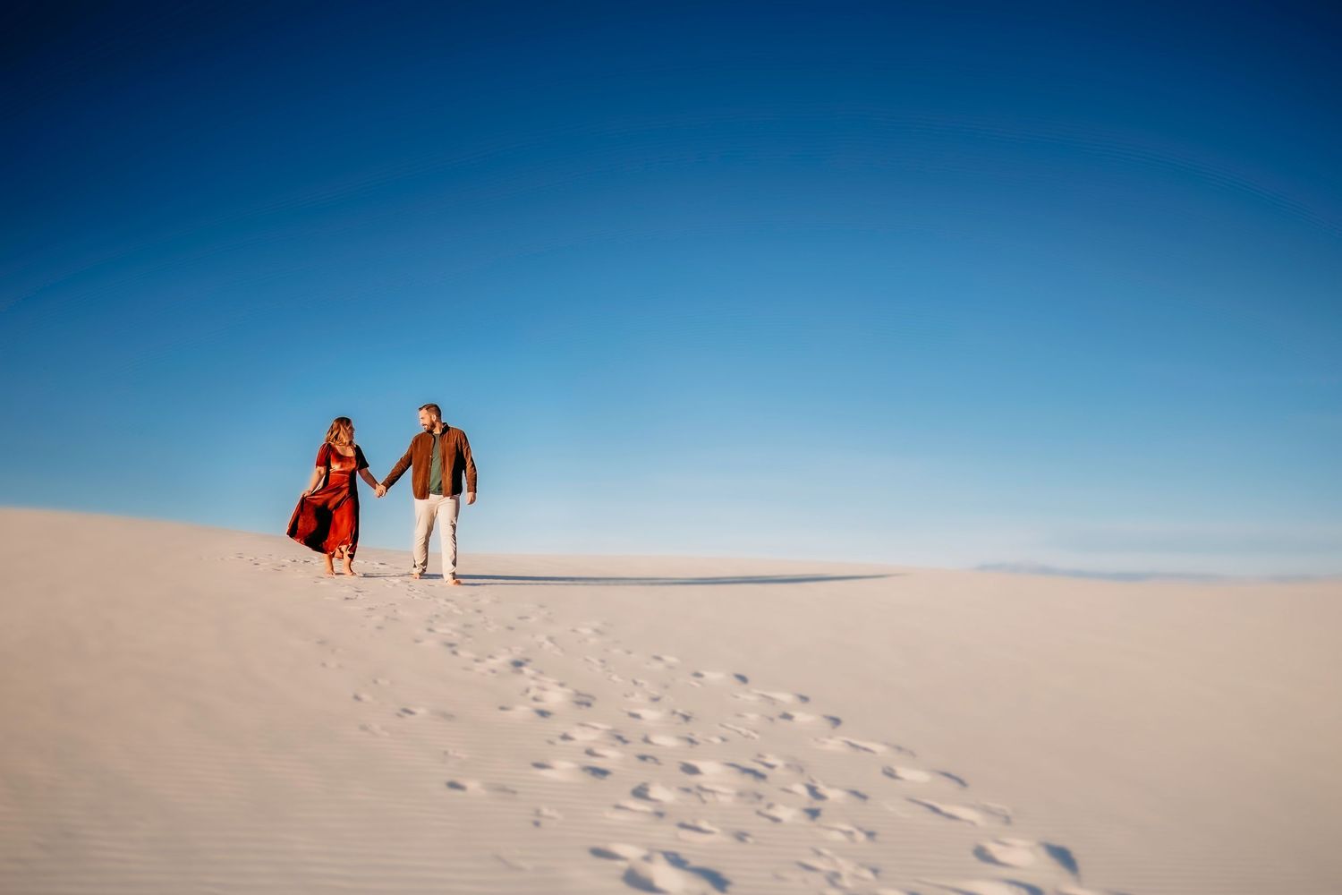 Couple holding hands walking on white sand dunes under a clear blue sky, woman in red dress.