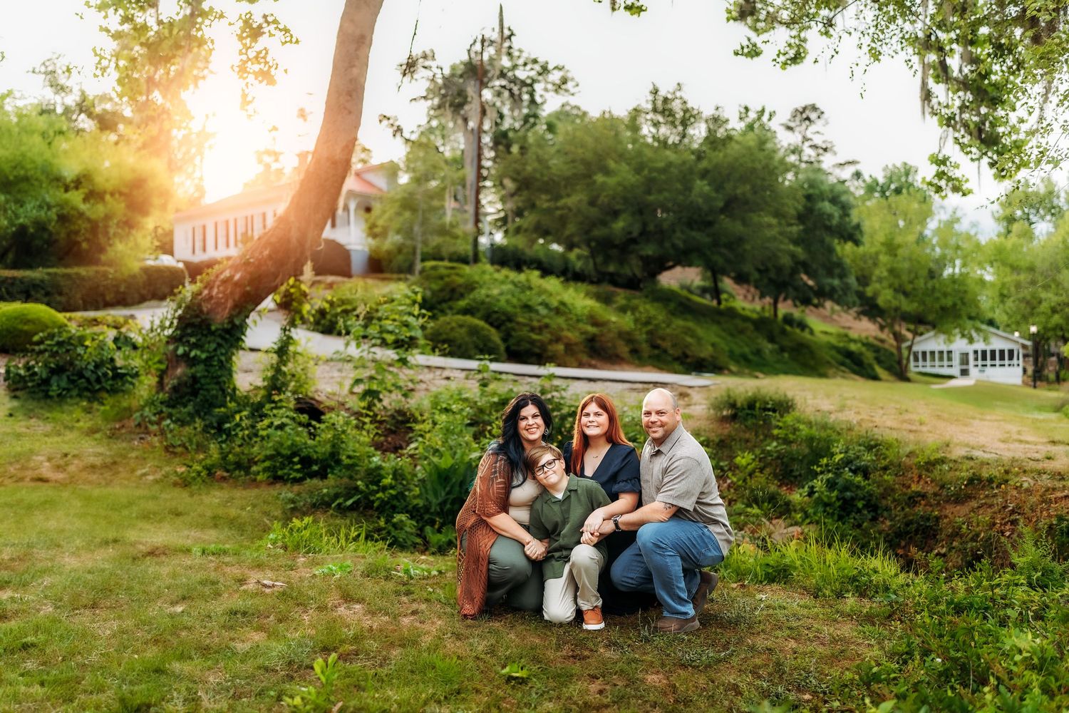 Family of four posing together on green grass at golden hour with sunlit trees and garden in background.