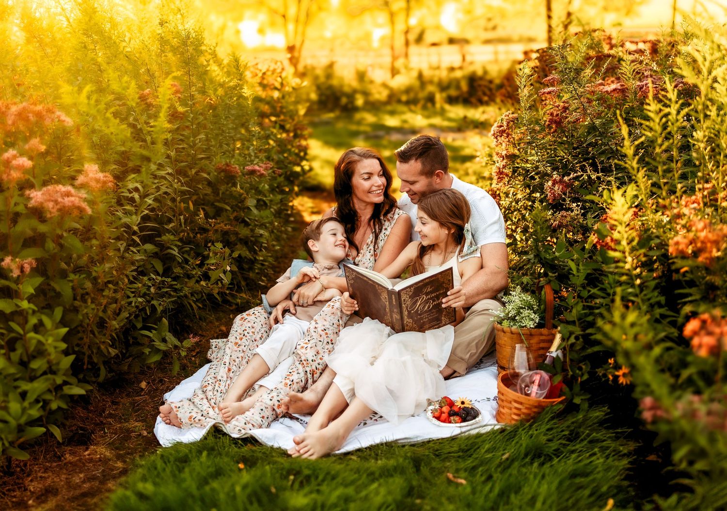 Family sitting on a blanket in a sunny garden, reading a book together surrounded by lush green bushes and flowers.