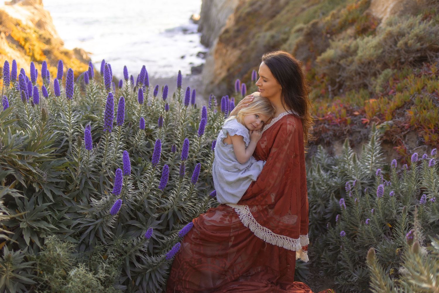Mother in red dress holds blonde toddler among purple wildflowers on a scenic coastal cliffside.