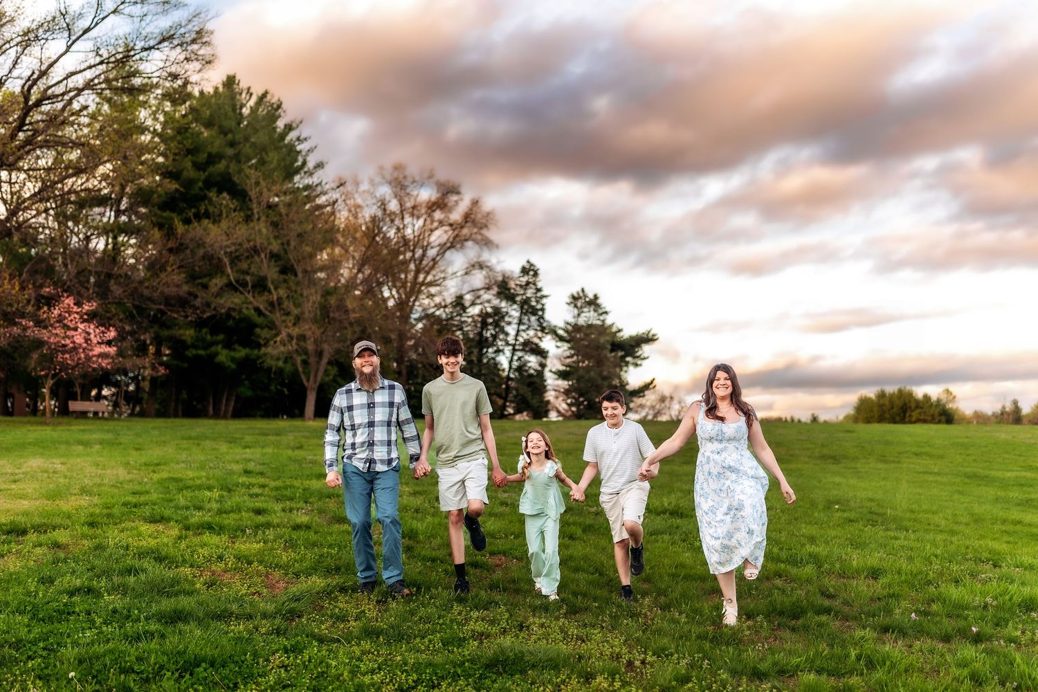 Family of five walking hand-in-hand across a green field at sunset with colorful clouds and trees in background.
