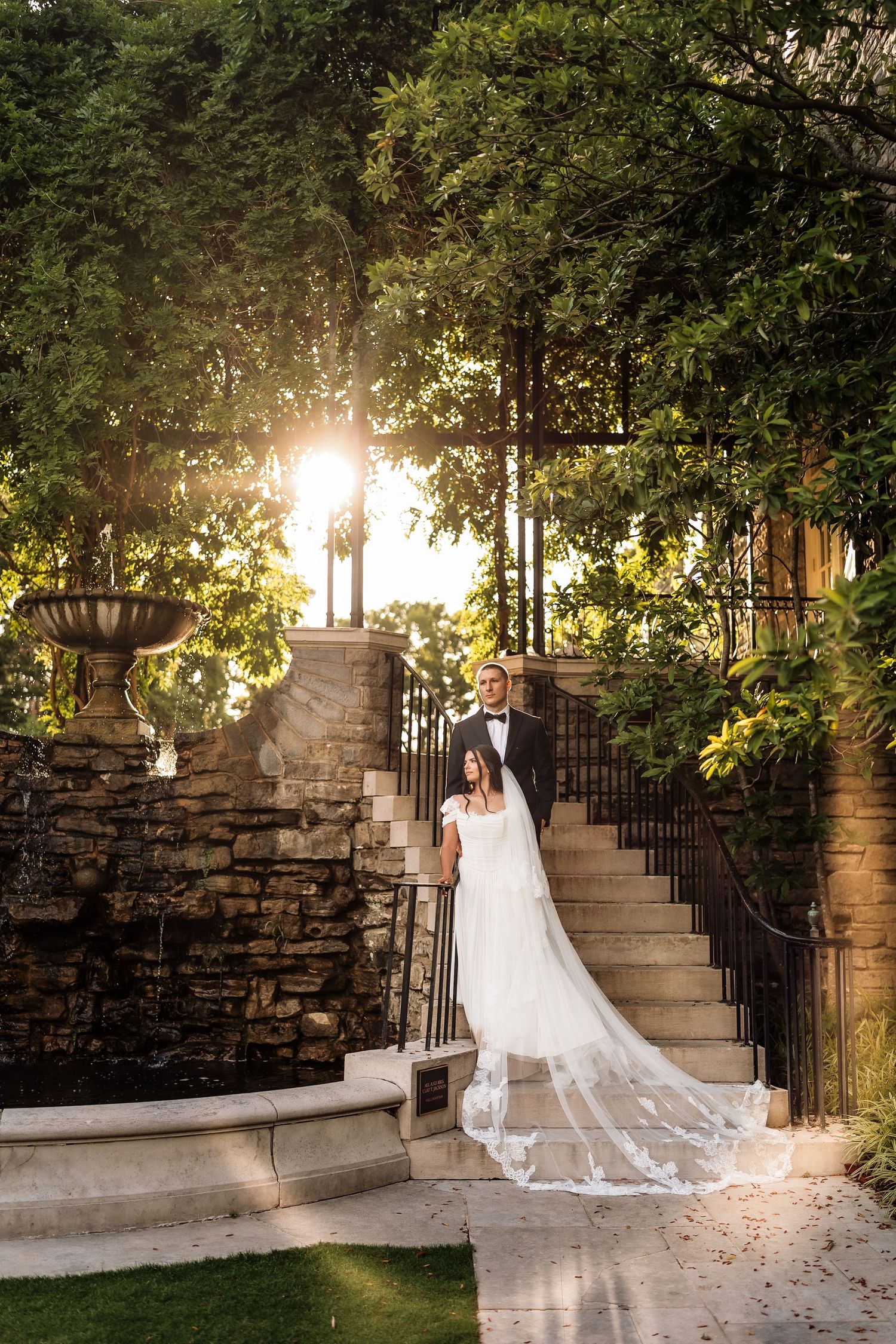 Bride and groom pose on stone steps surrounded by lush greenery with golden sunlight glowing behind them.