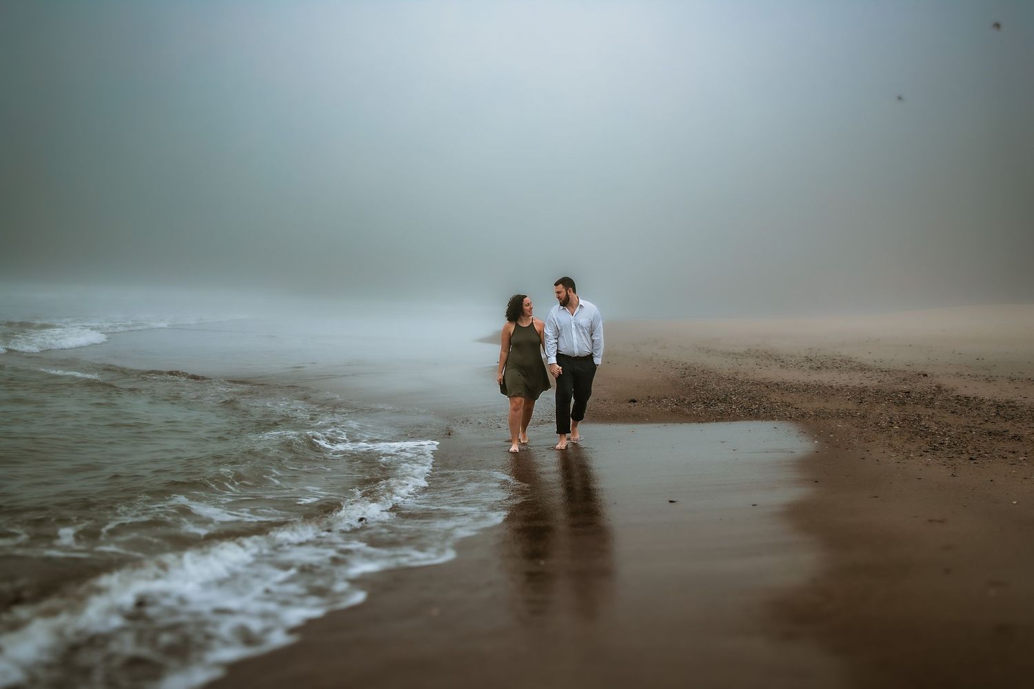 Couple walking hand in hand along a misty beach shoreline, waves gently lapping at their feet on a foggy day.