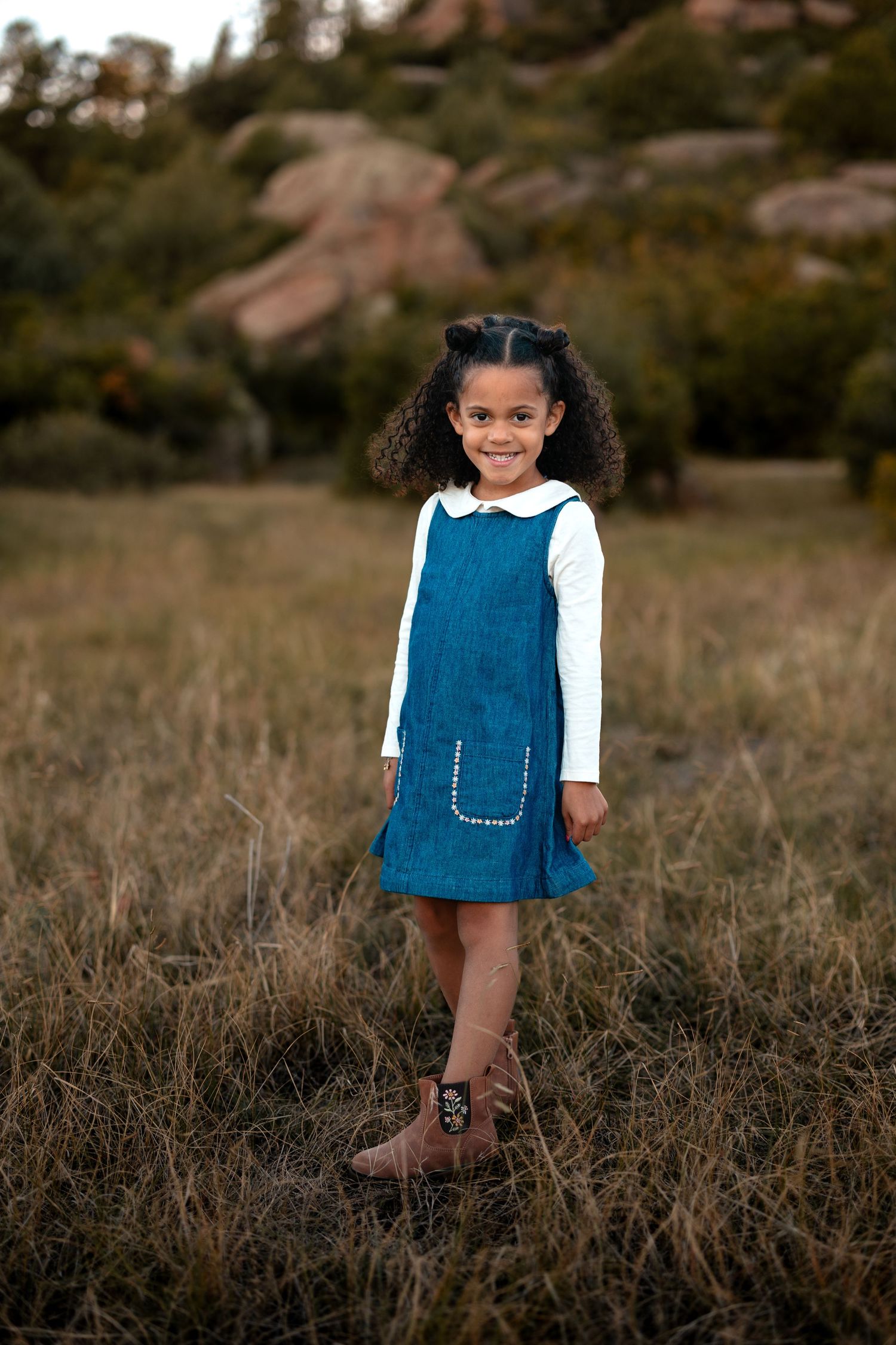 Smiling young girl with curly pigtails in a blue denim pinafore and brown boots standing in a golden grass field with rocky hills behind.