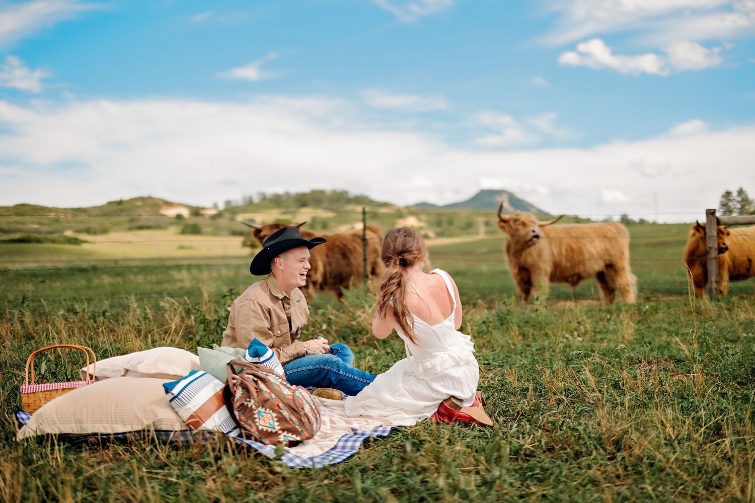 Couple enjoying a picnic in a green meadow with highland cattle grazing nearby under a partly cloudy sky.