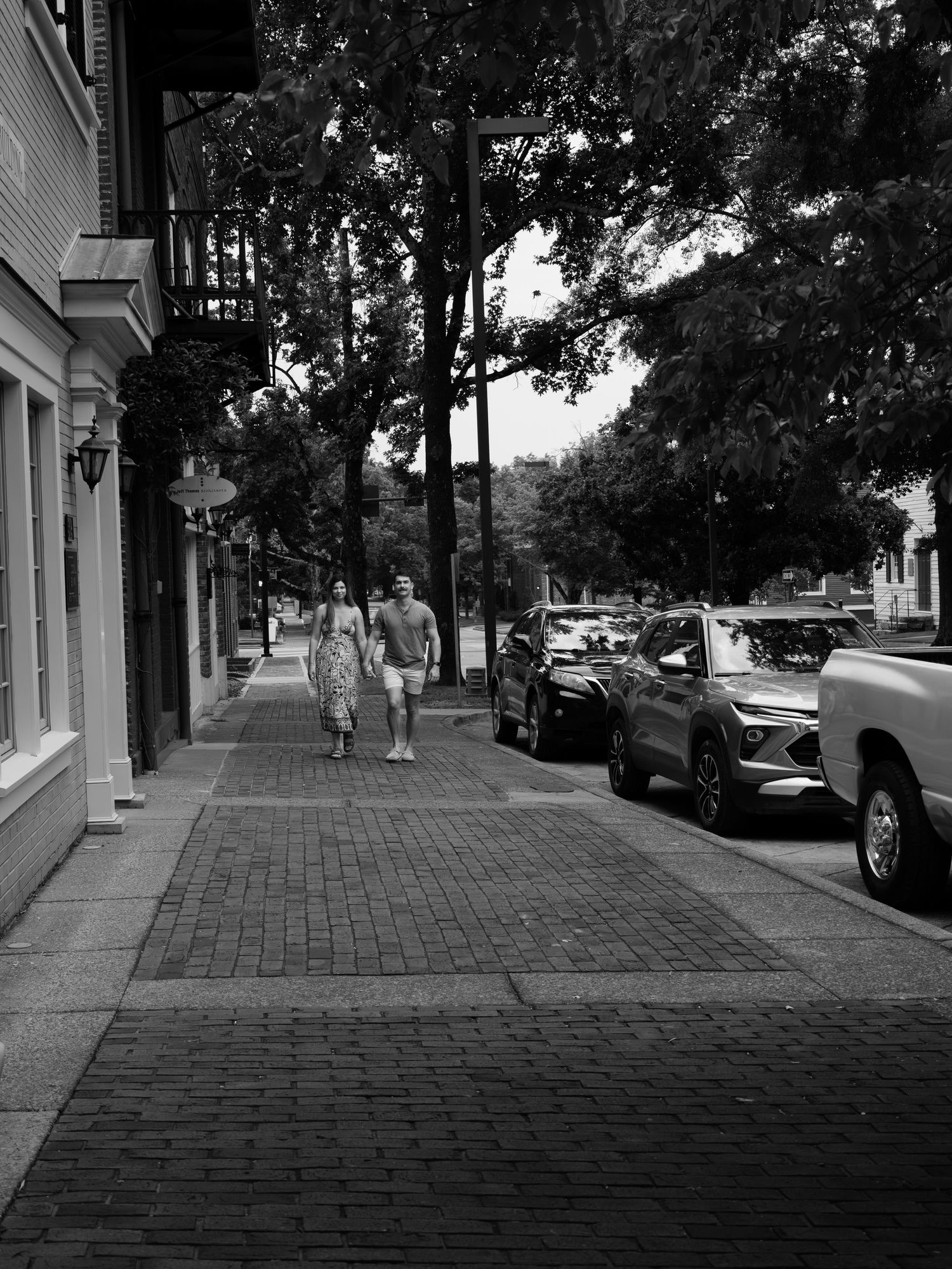 Black and white photo of two people walking on a brick sidewalk lined with trees and parked cars in a small town.
