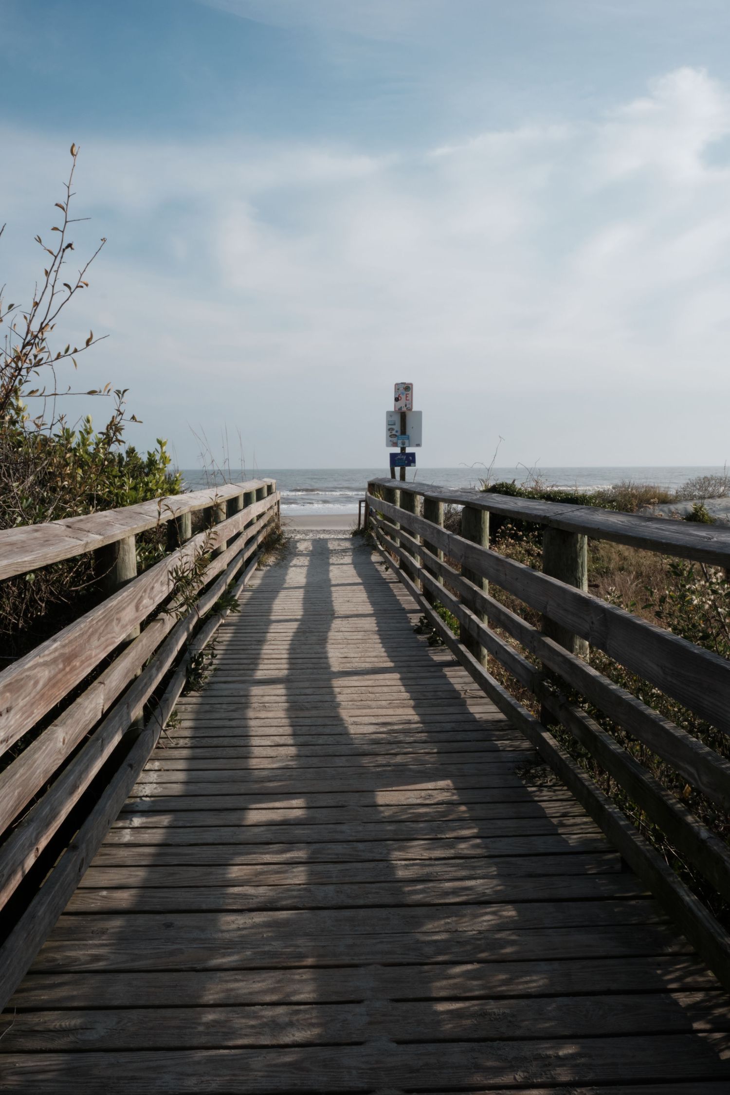 Wooden boardwalk pathway leading toward the ocean beach under a partly cloudy blue sky.
