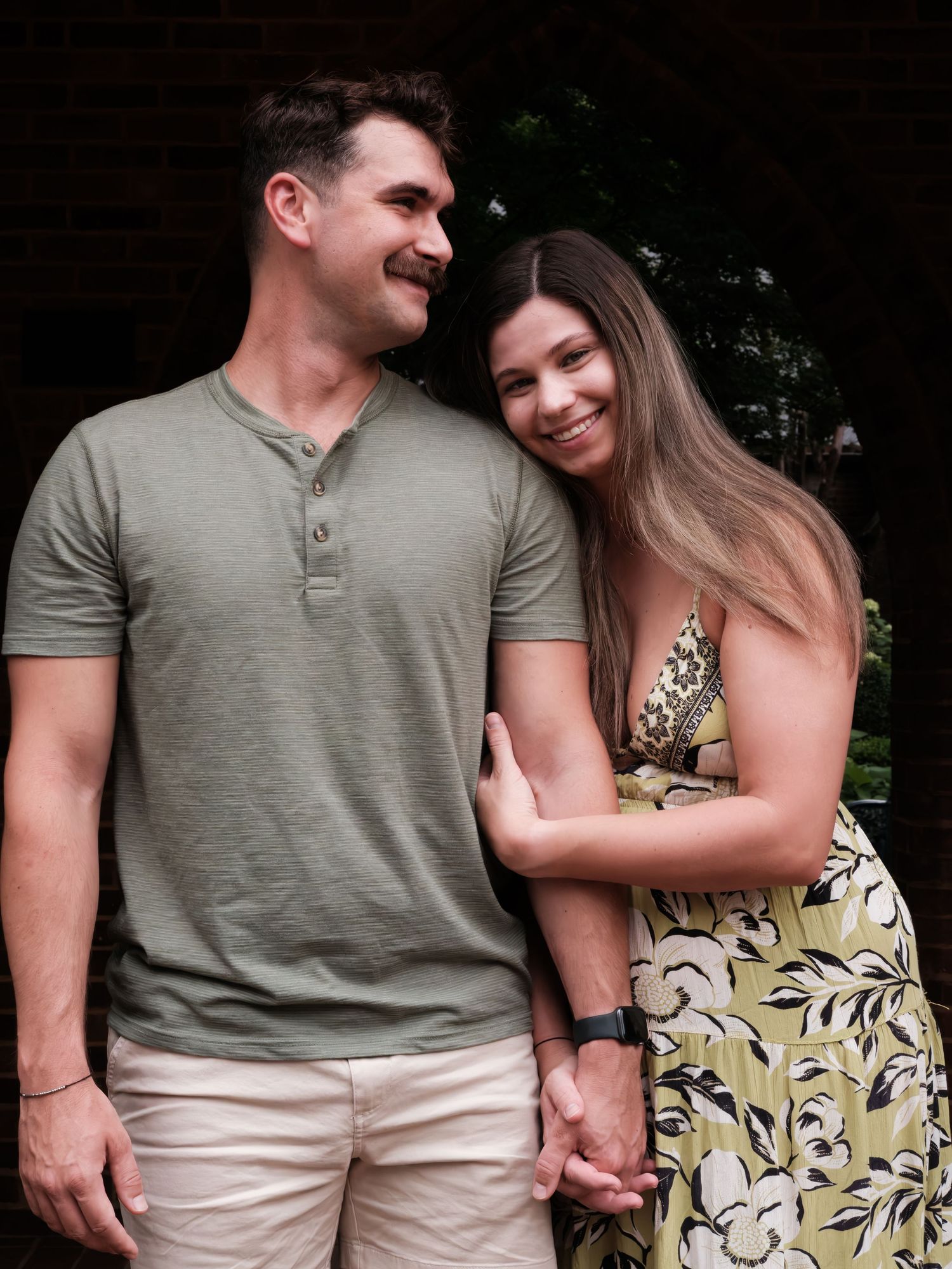 Happy couple posing outdoors, man in olive henley and woman in floral maxi dress smiling warmly together.