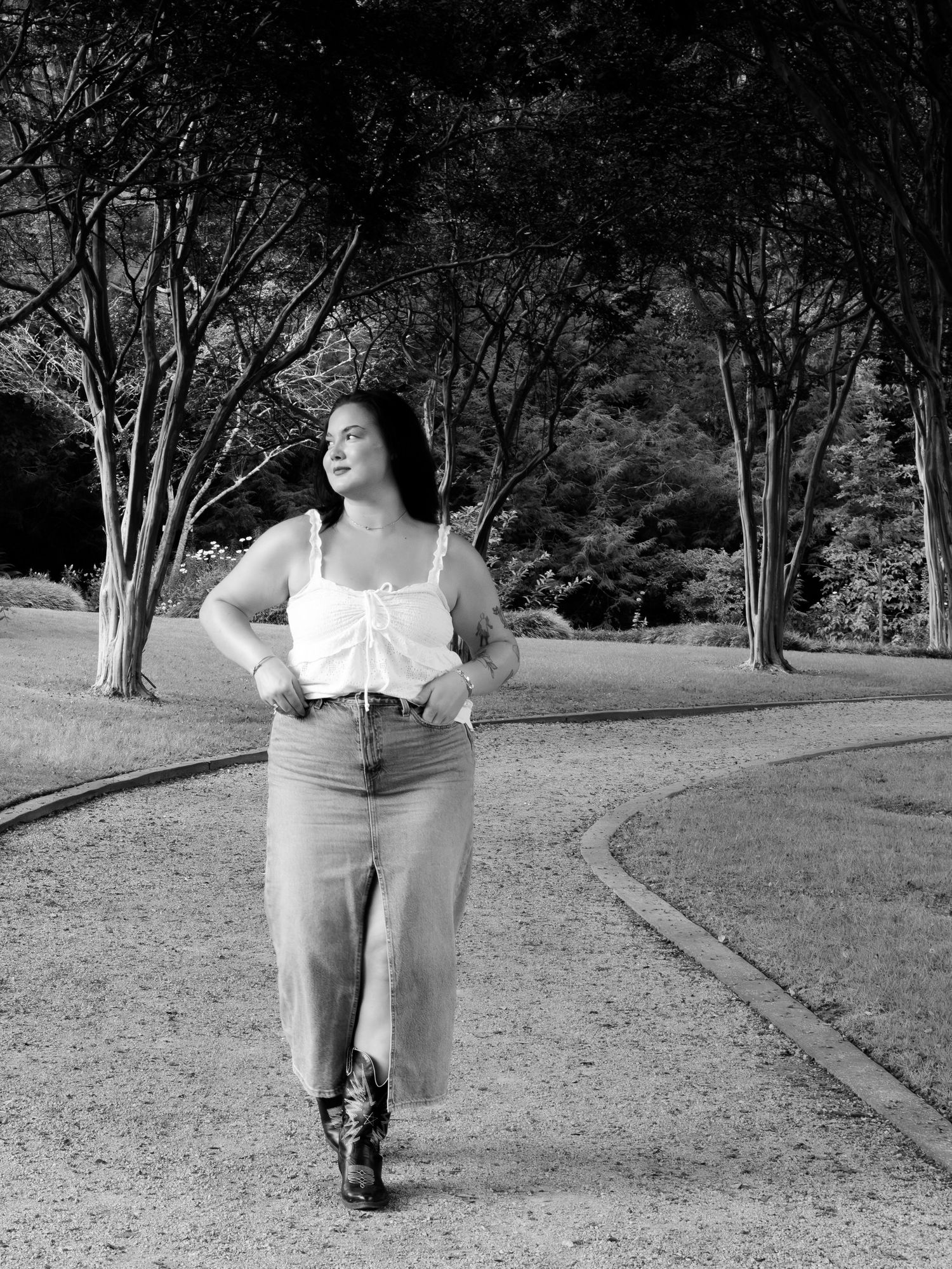 Black and white photo of a woman in a white top and denim skirt walking on a park path.