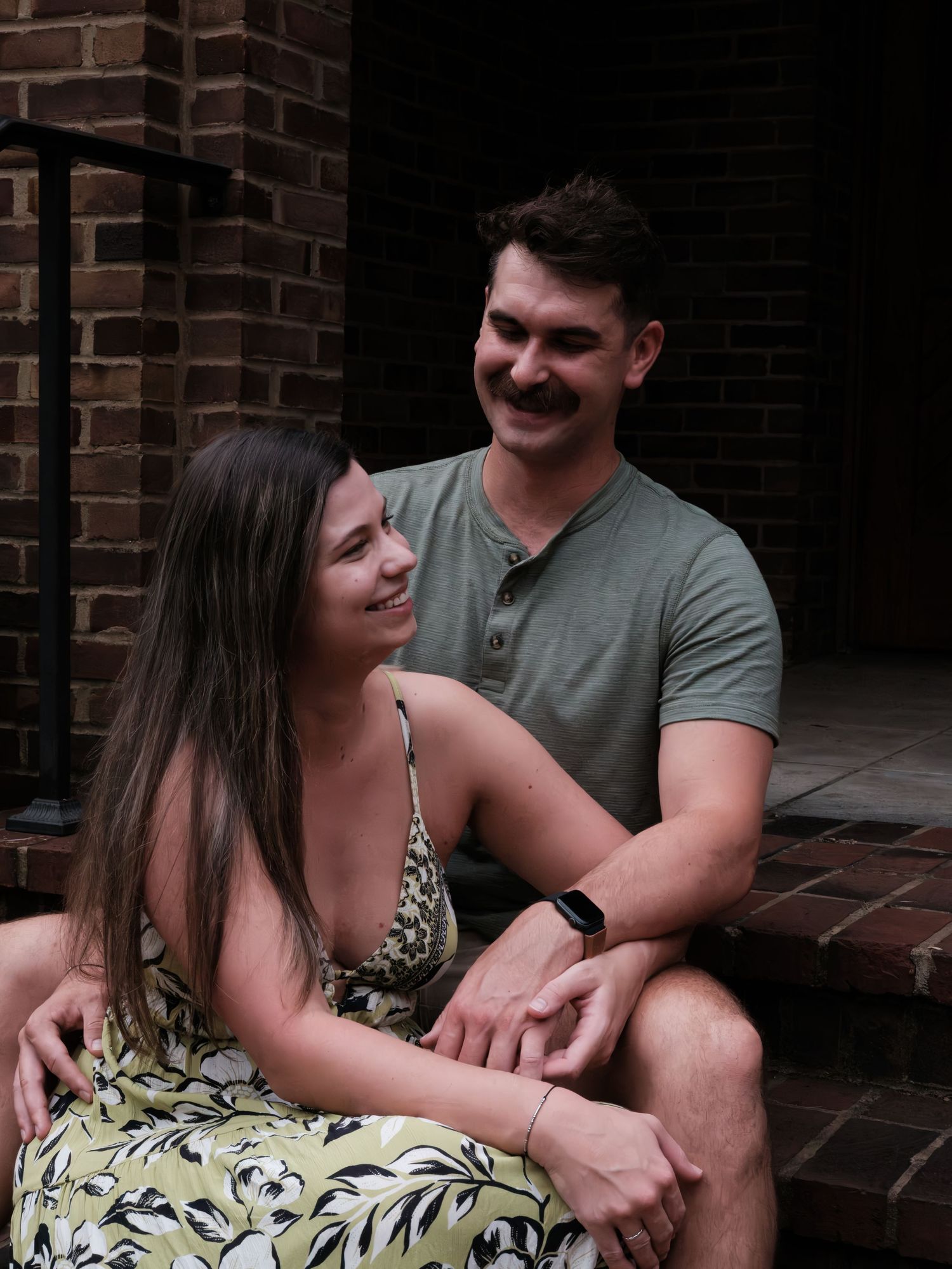 Happy couple sitting on brick steps, smiling at each other, woman in floral dress, man in green henley shirt.