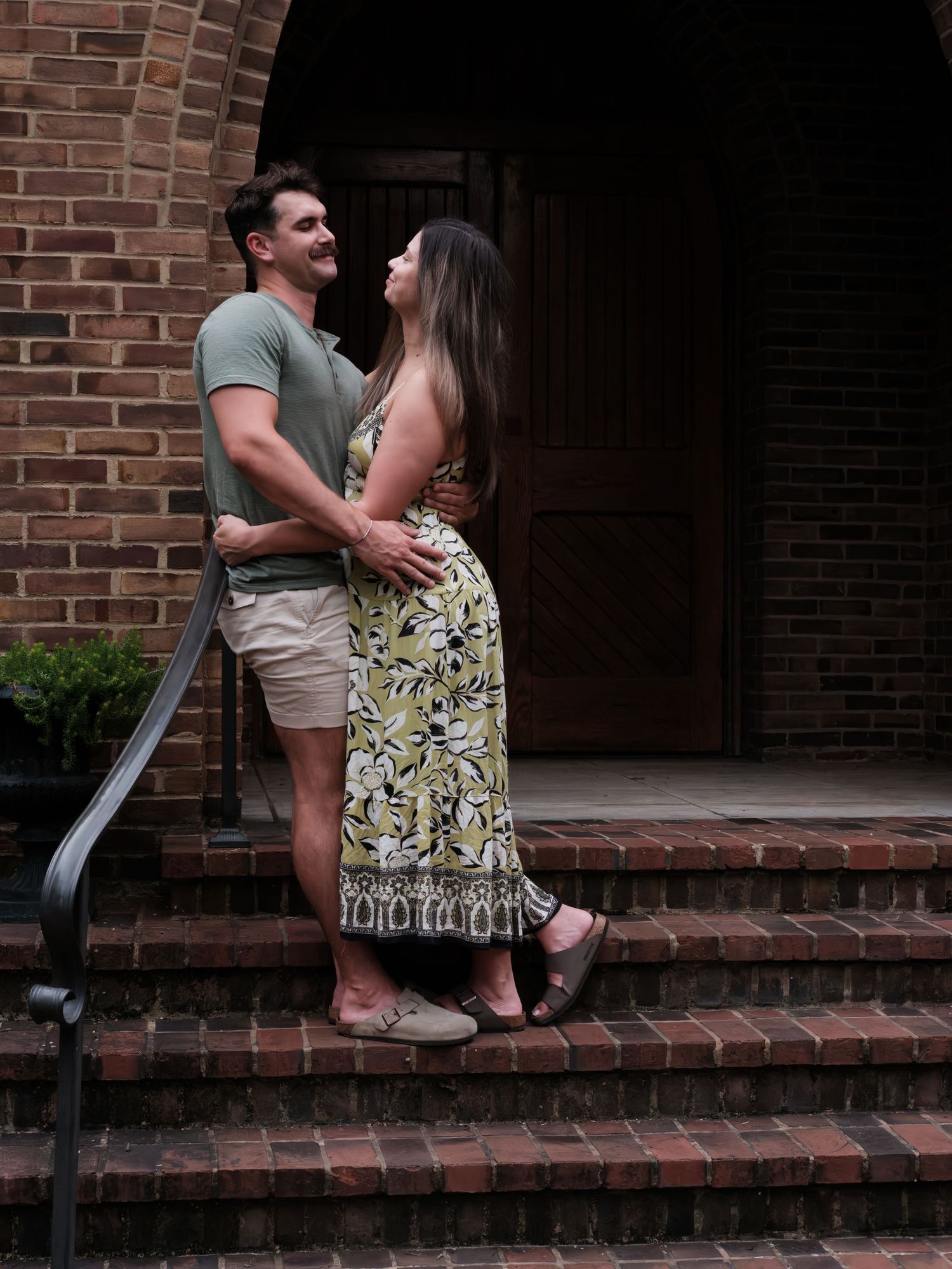 Romantic couple embracing on brick steps of a historic building, woman in yellow floral maxi dress, man in green shirt.