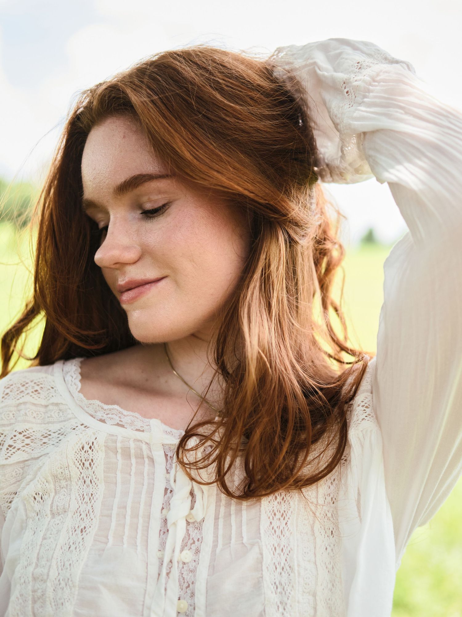 Young redhead woman in a white lace blouse poses outdoors in a sunlit open field.
