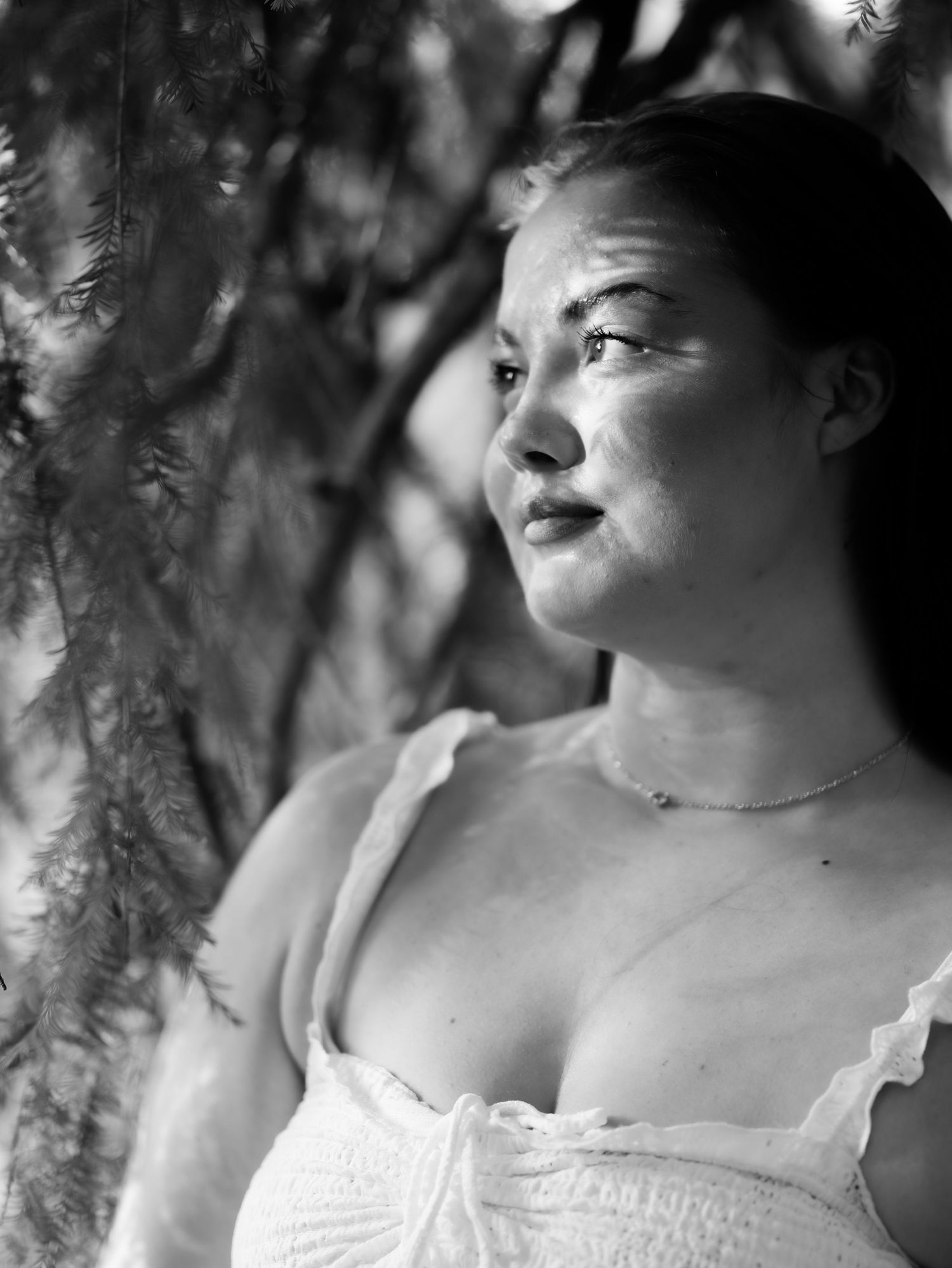 Black and white portrait of a woman gazing upward beside hanging tree branches in natural light.
