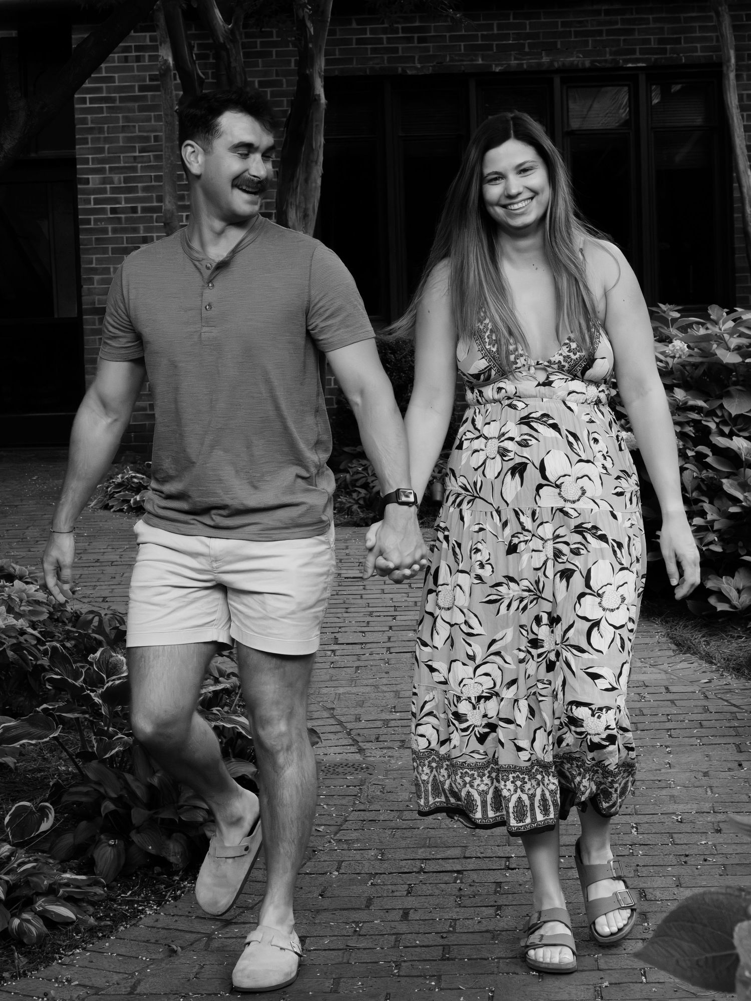 Black and white photo of a happy couple holding hands and walking on a brick pathway outdoors.