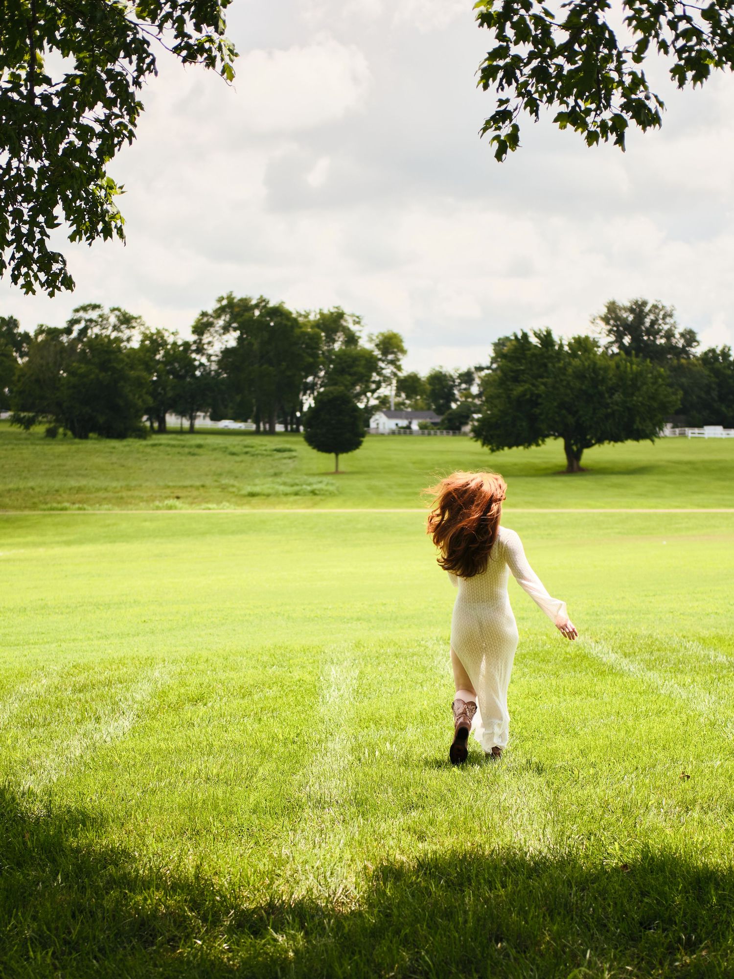 Redhead woman in a white dress runs across a lush green lawn surrounded by trees.