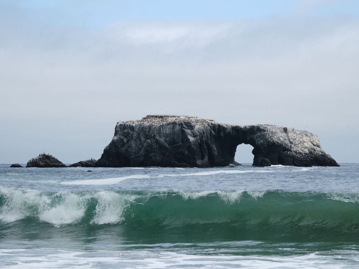 Natural rock arch formation rising from turquoise ocean waves under overcast sky at California coastline.