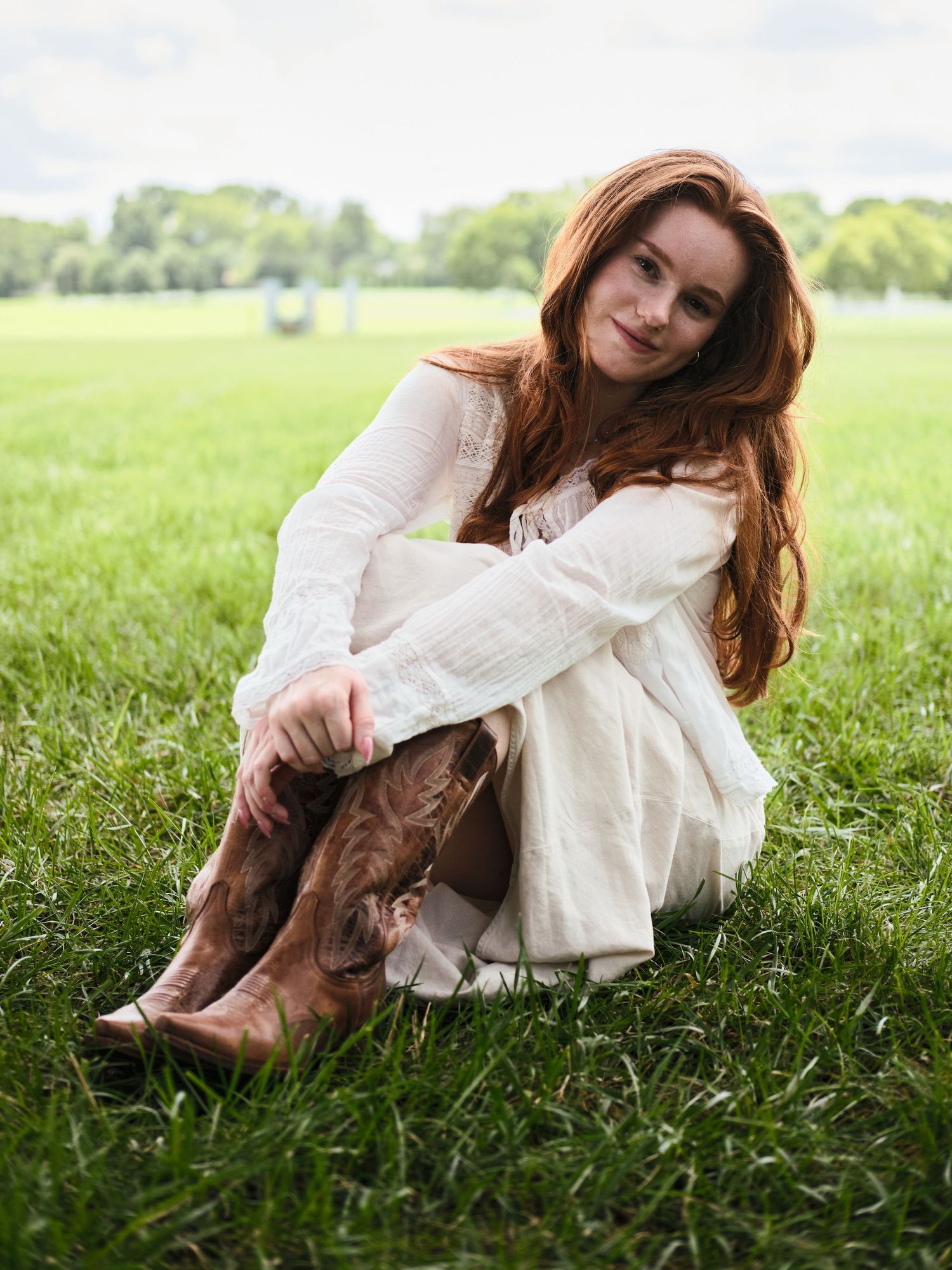 Red-haired woman sitting cross-legged in lush green grass wearing a white linen dress and brown cowboy boots.