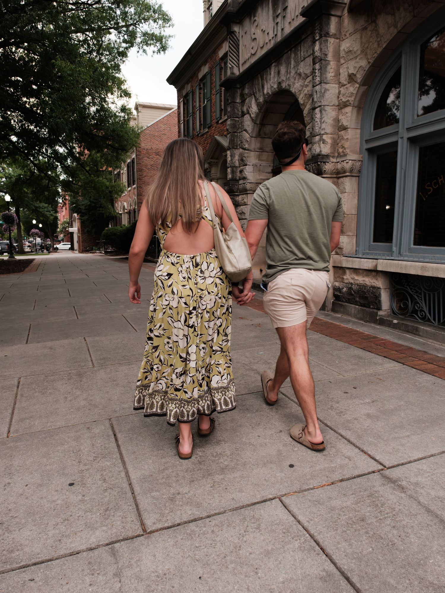 Couple strolling hand in hand down a charming urban sidewalk past a historic stone building.