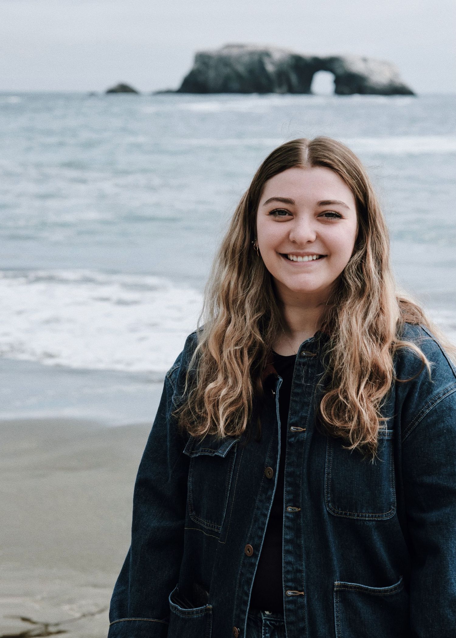 Smiling young woman in denim jacket poses on beach with natural rock arch formation in ocean behind her.