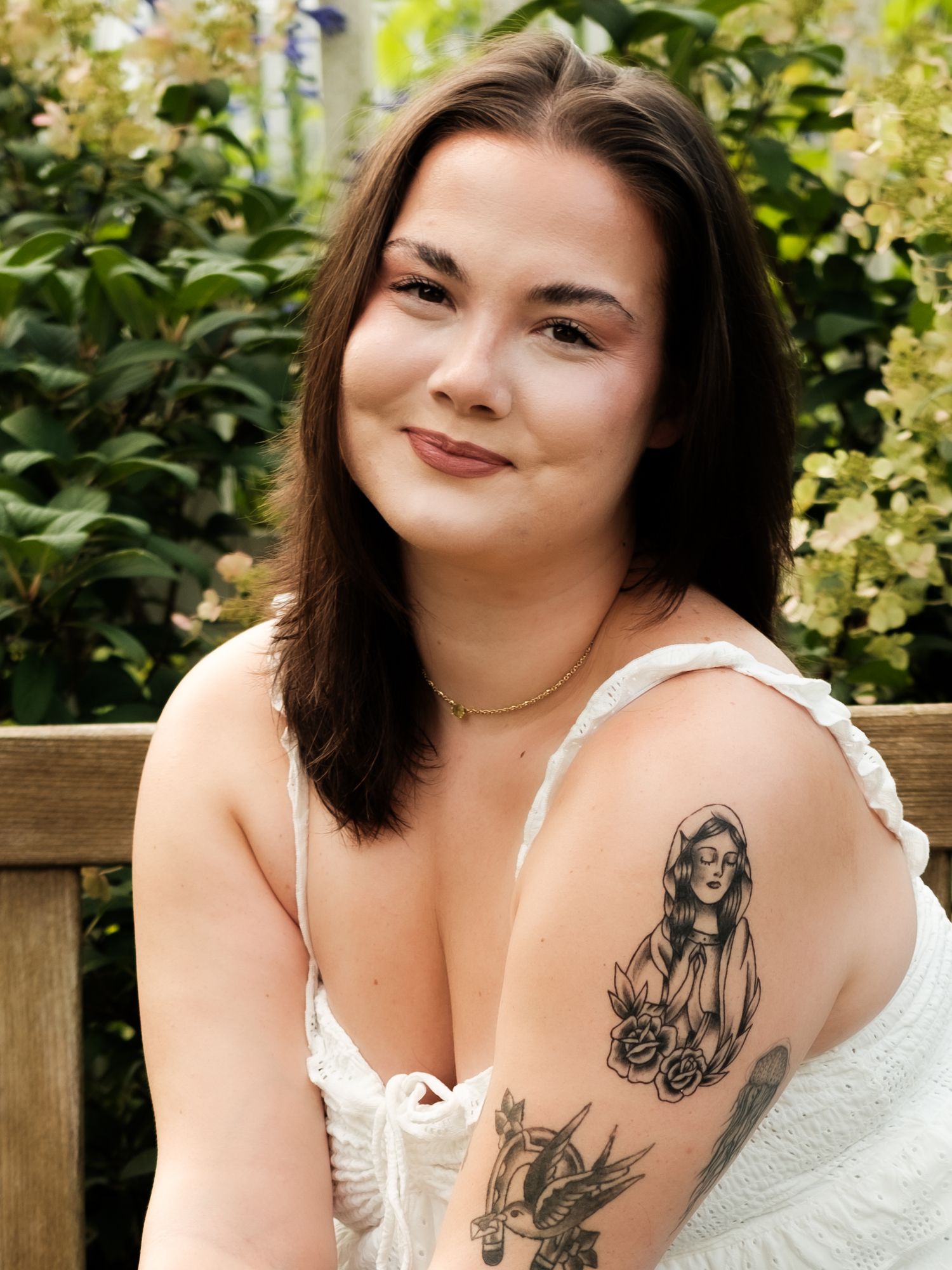 Smiling brunette woman with arm tattoos sits outdoors in front of blooming green shrubs.