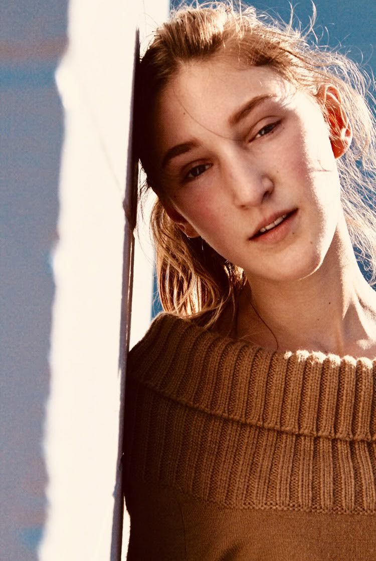 A young woman in a tan knit sweater leans against a white wall in warm sunlight.