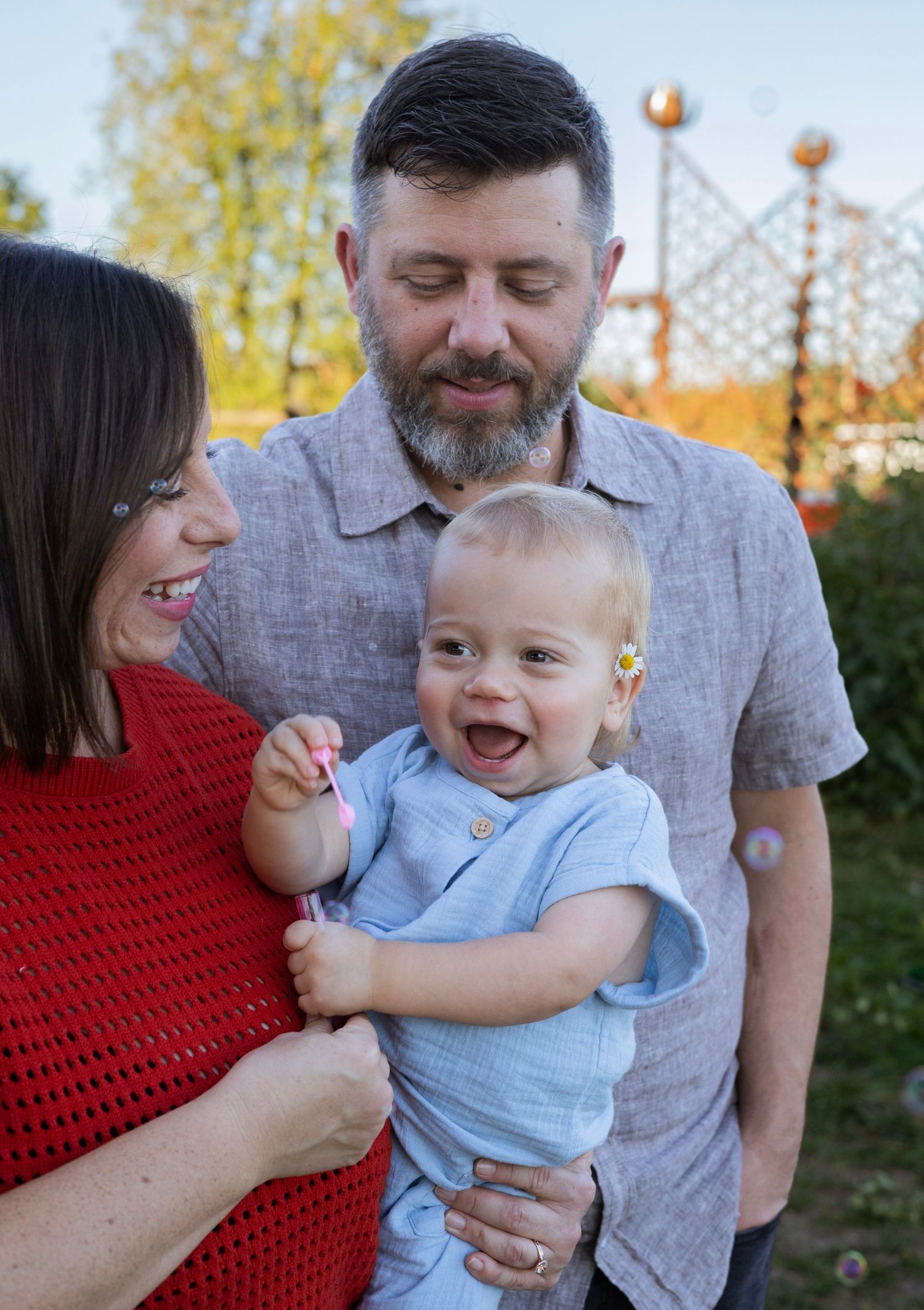 Happy family portrait with smiling baby, loving mother in red, and bearded father outdoors in a sunny park.