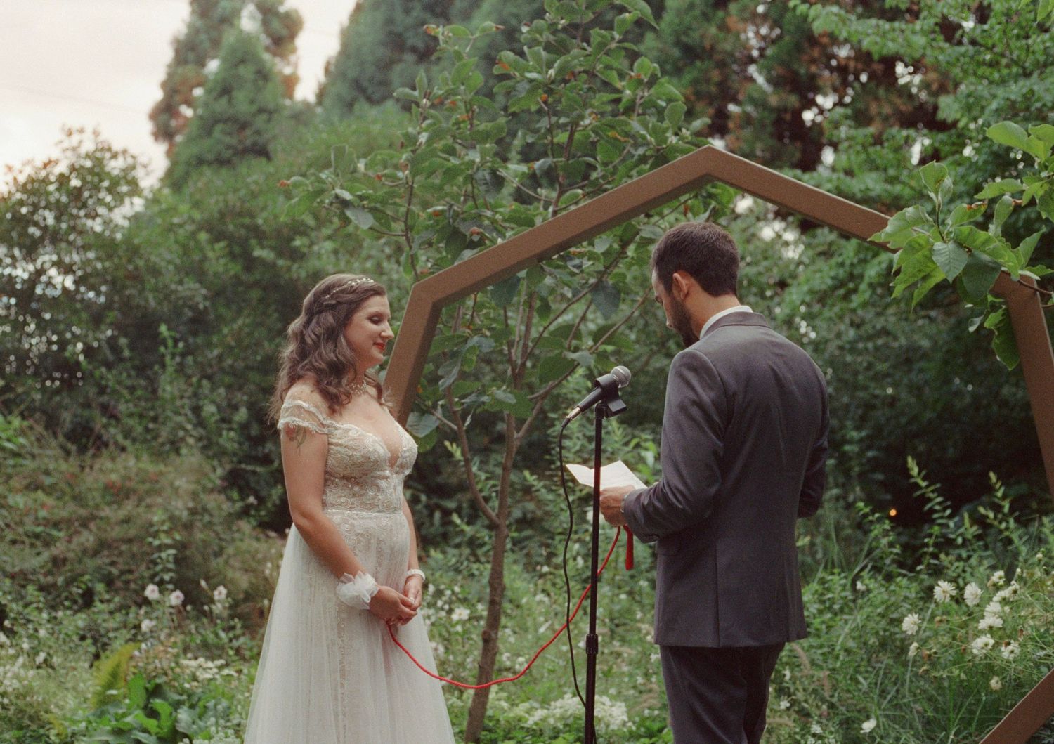 Bride and groom exchange vows outdoors under a geometric wooden arch surrounded by lush greenery.