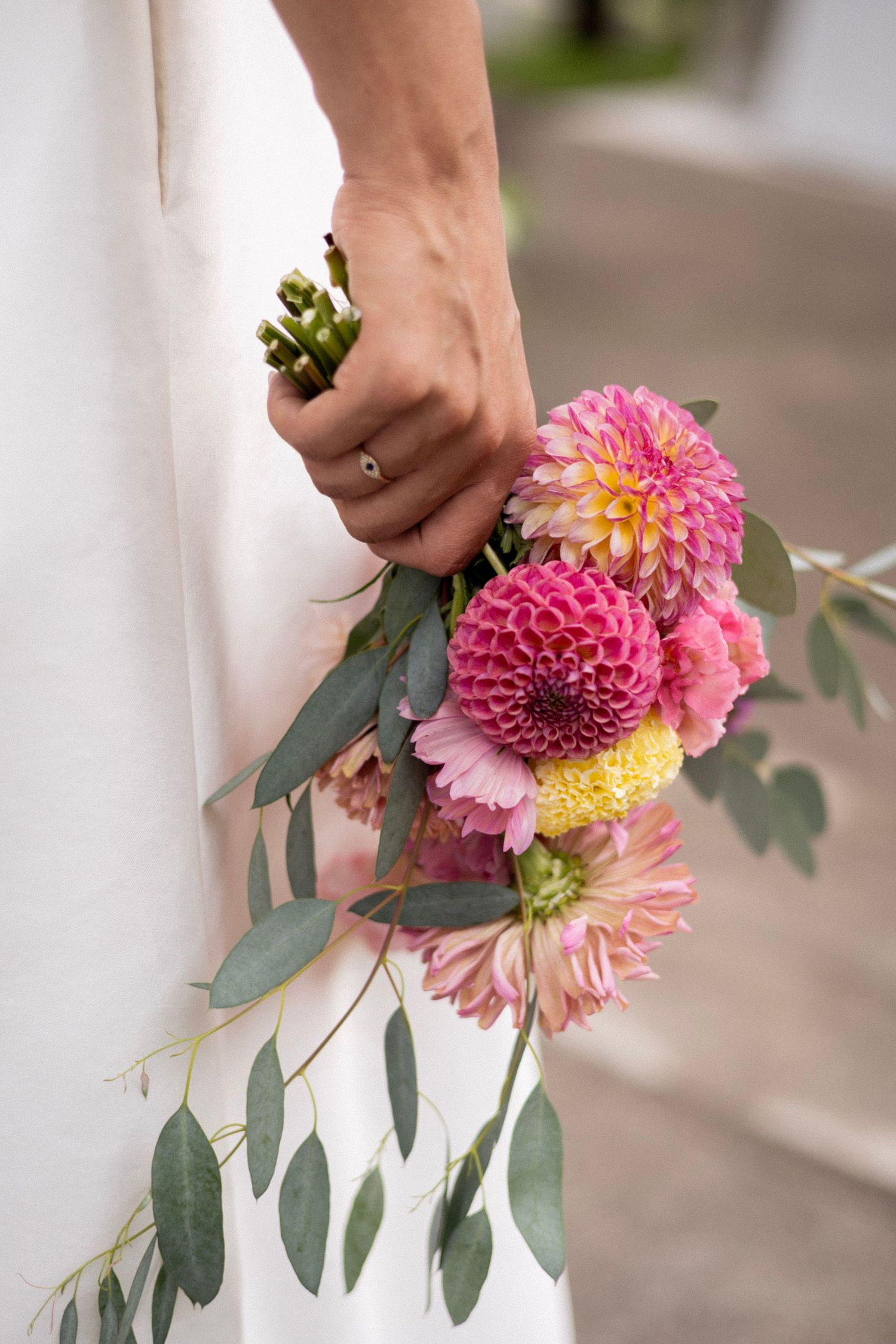 Bride holding vibrant pink and yellow dahlia wedding bouquet with eucalyptus leaves against white gown.