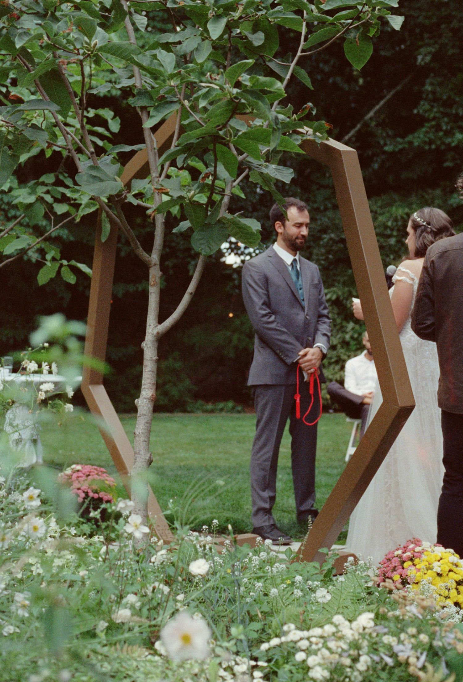 Groom in gray suit stands under geometric hexagonal wooden arch during outdoor garden wedding ceremony.